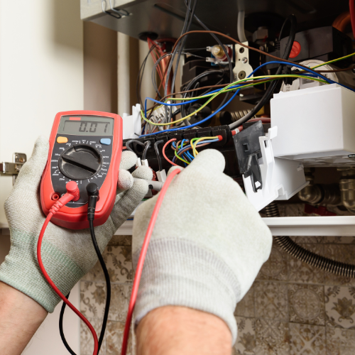 Person wearing gloves using a digital multimeter to test wiring inside an electrical panel with wires and components. Norwich Plumbing and Heating Ltd.