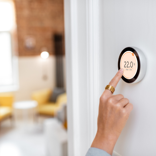Close-up of a person's finger adjusting a round smart thermostat on a wall, displaying 22.0 degrees Celsius, in a modern living room with yellow chairs and a brick wall in the background. Norwich Plumbing and Heating Ltd.