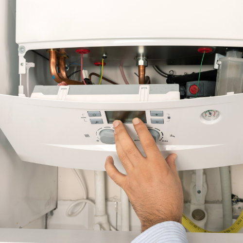Close-up of a person's hand adjusting a thermostat or control panel on a wall-mounted boiler or water heater with visible internal pipes and components. Norwich Plumbing and Heating Ltd.