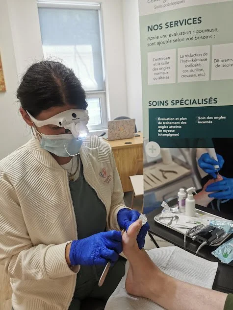 Une professionnelle de la santé, portant une blouse blanche, un masque et des gants bleus, examine un pied avec des outils médico-dentaires dans un cabinet médical, à côté d'une affiche informative.