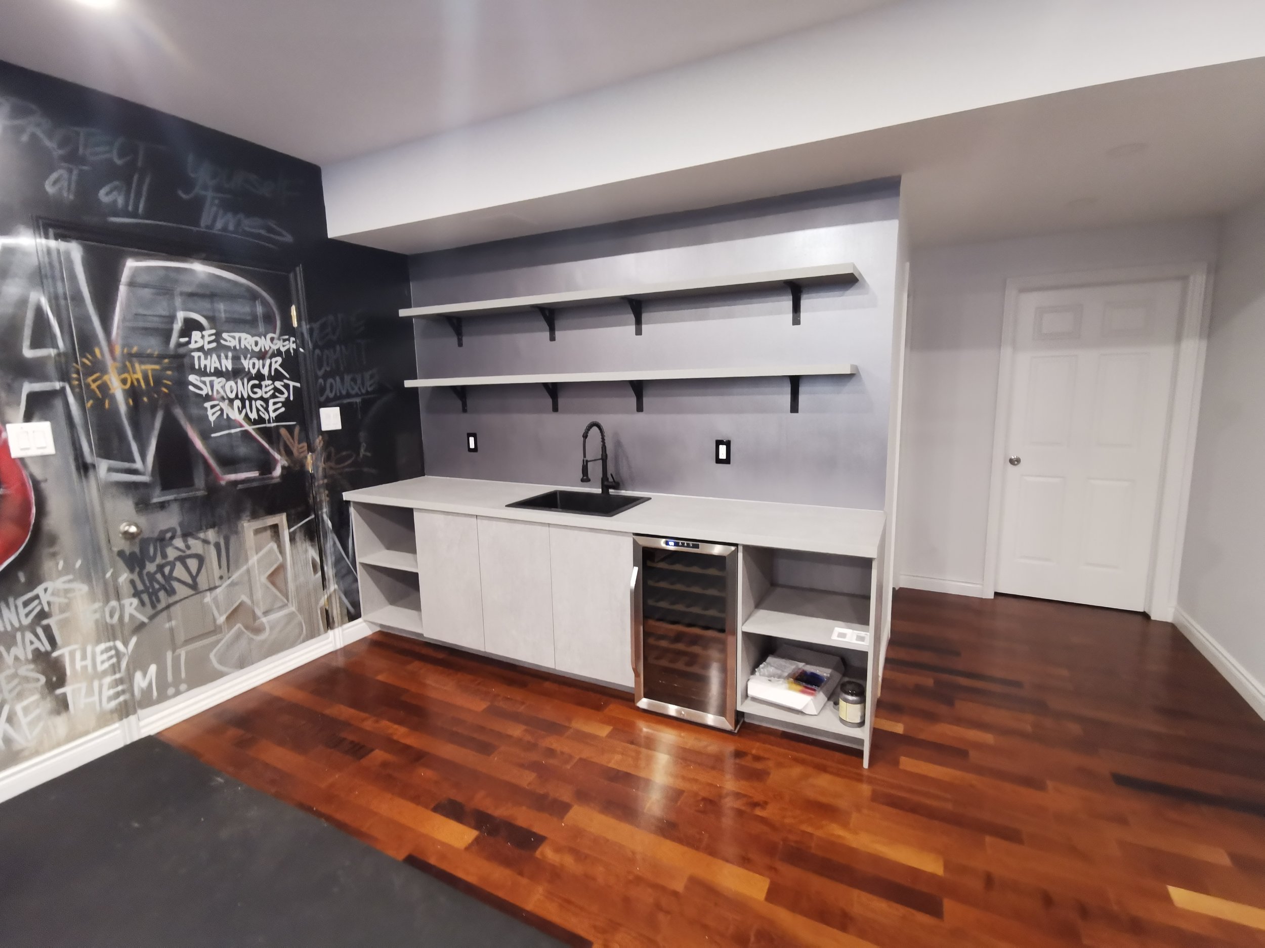 A small kitchenette with floating shelves, a black sink, and a wine cooler, with a black chalkboard wall featuring motivational words and drawings to the left and a white door to the right. The floor is dark hardwood.