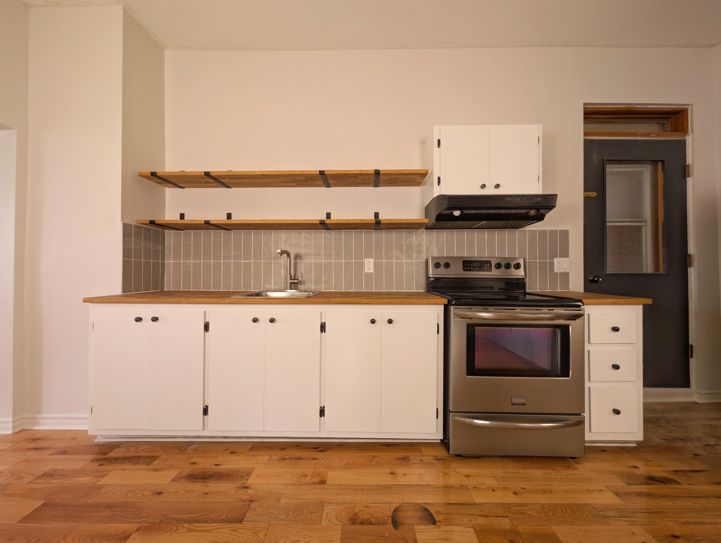 A minimalist kitchen with white cabinets, a stainless steel stove, a wooden countertop, open wooden shelves, and a gray tiled backsplash, with a black door on the right.