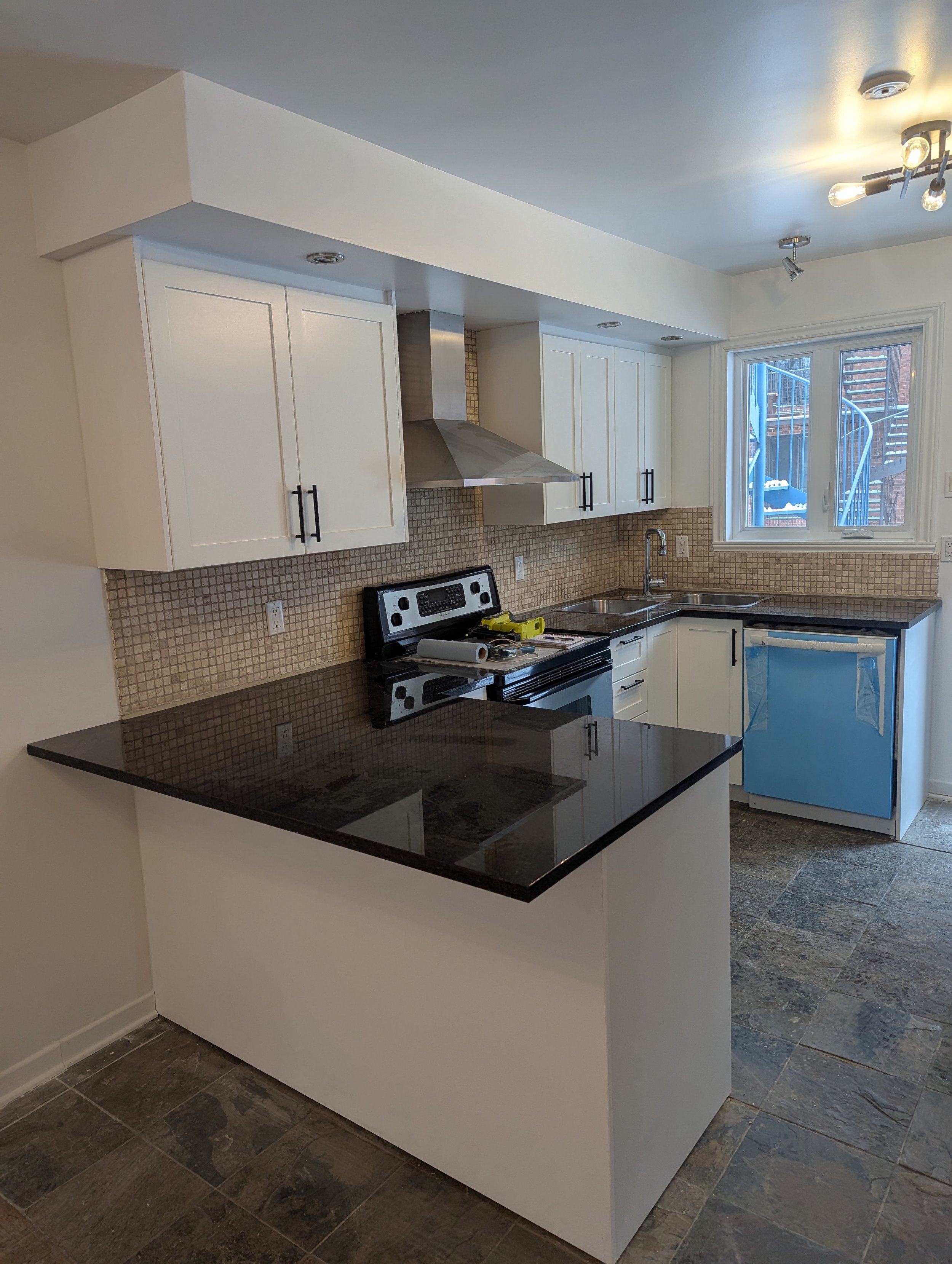 Modern kitchen with white cabinets, black countertop, stainless steel range hood, and a window overlooking an outdoor staircase.