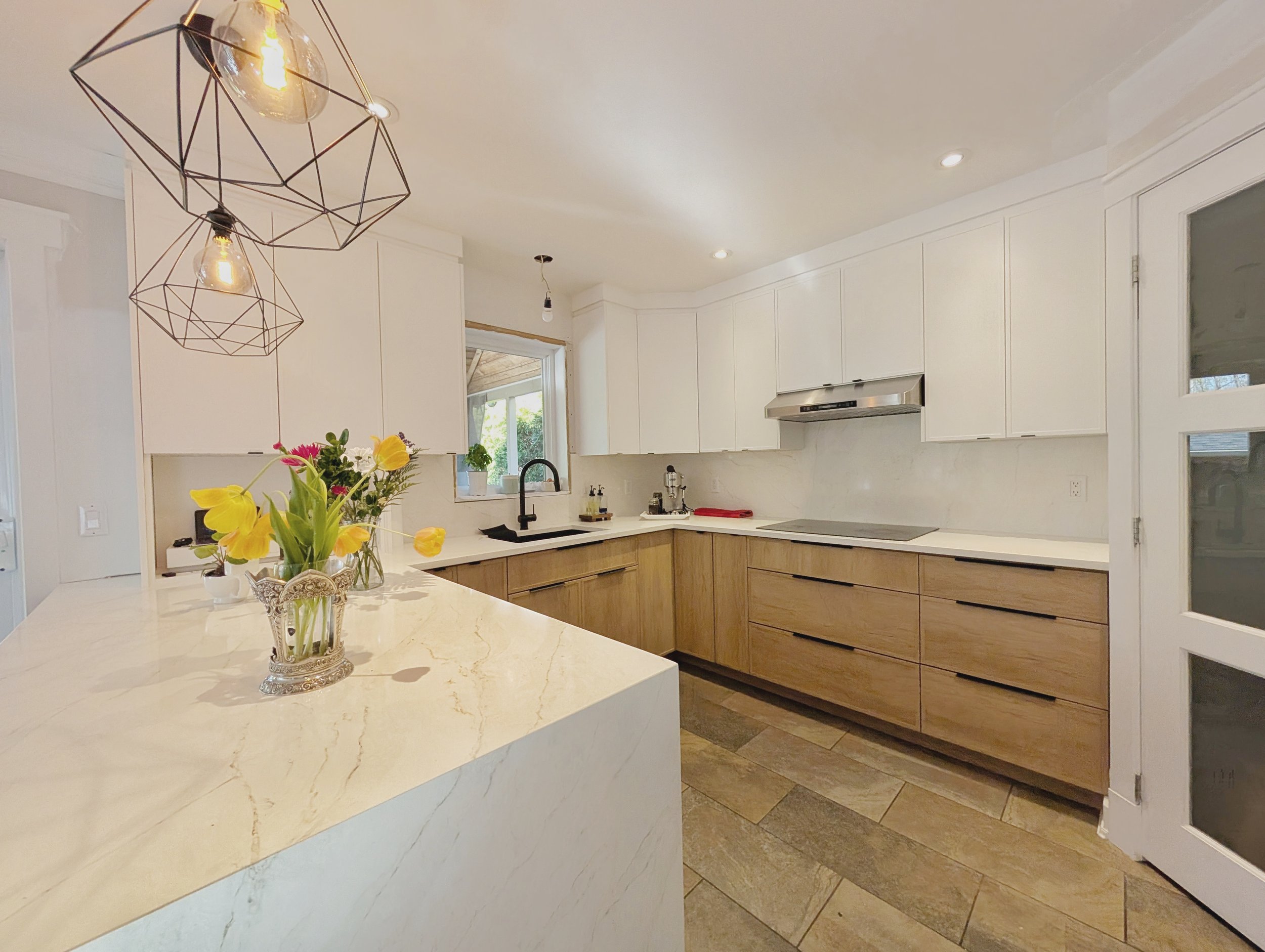 Modern kitchen with white and wood cabinetry, marble countertops, a faucet sink under a window, decorative pendant lights, a vase with yellow tulips on an island, and a tiled floor.