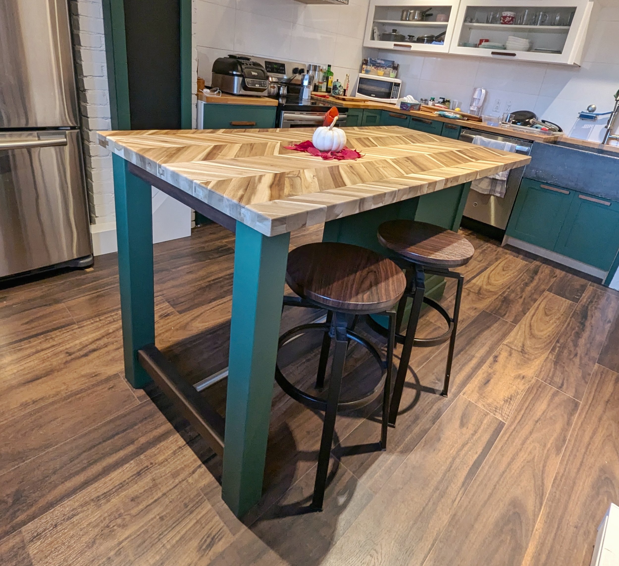 Kitchen with a wooden island table, green base, two black bar stools, a pumpkin centerpiece, and various kitchen appliances and open cabinets in the background.