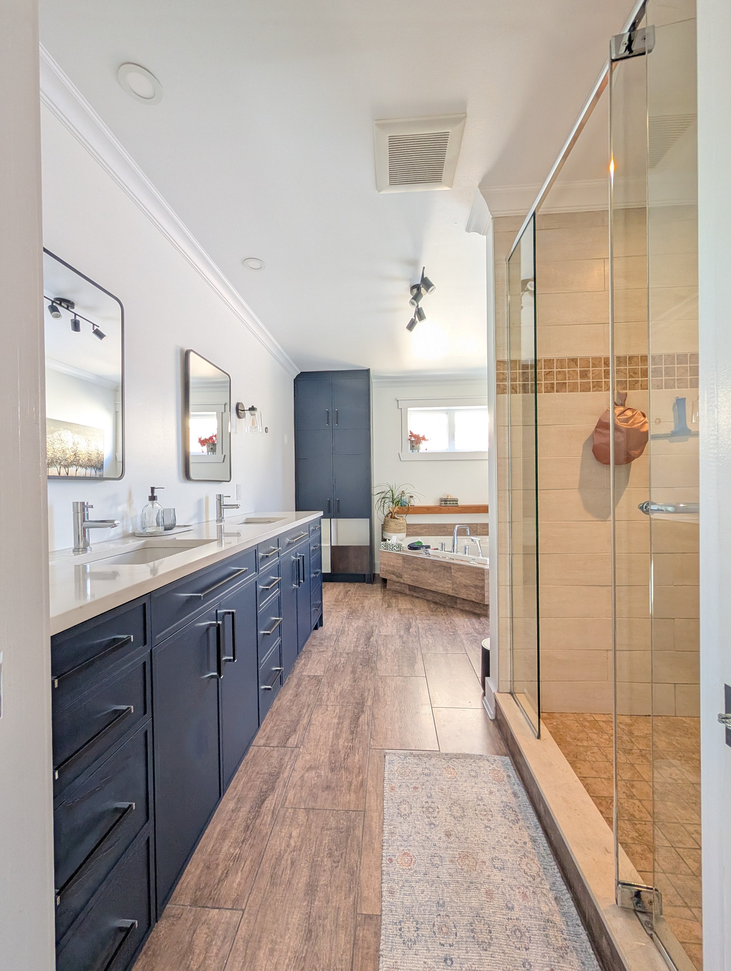 Modern bathroom with dual sinks, navy blue cabinets, wall mirrors, a large shower with glass doors, and a bathtub in the corner. The room has wood floor, small window, and white walls.