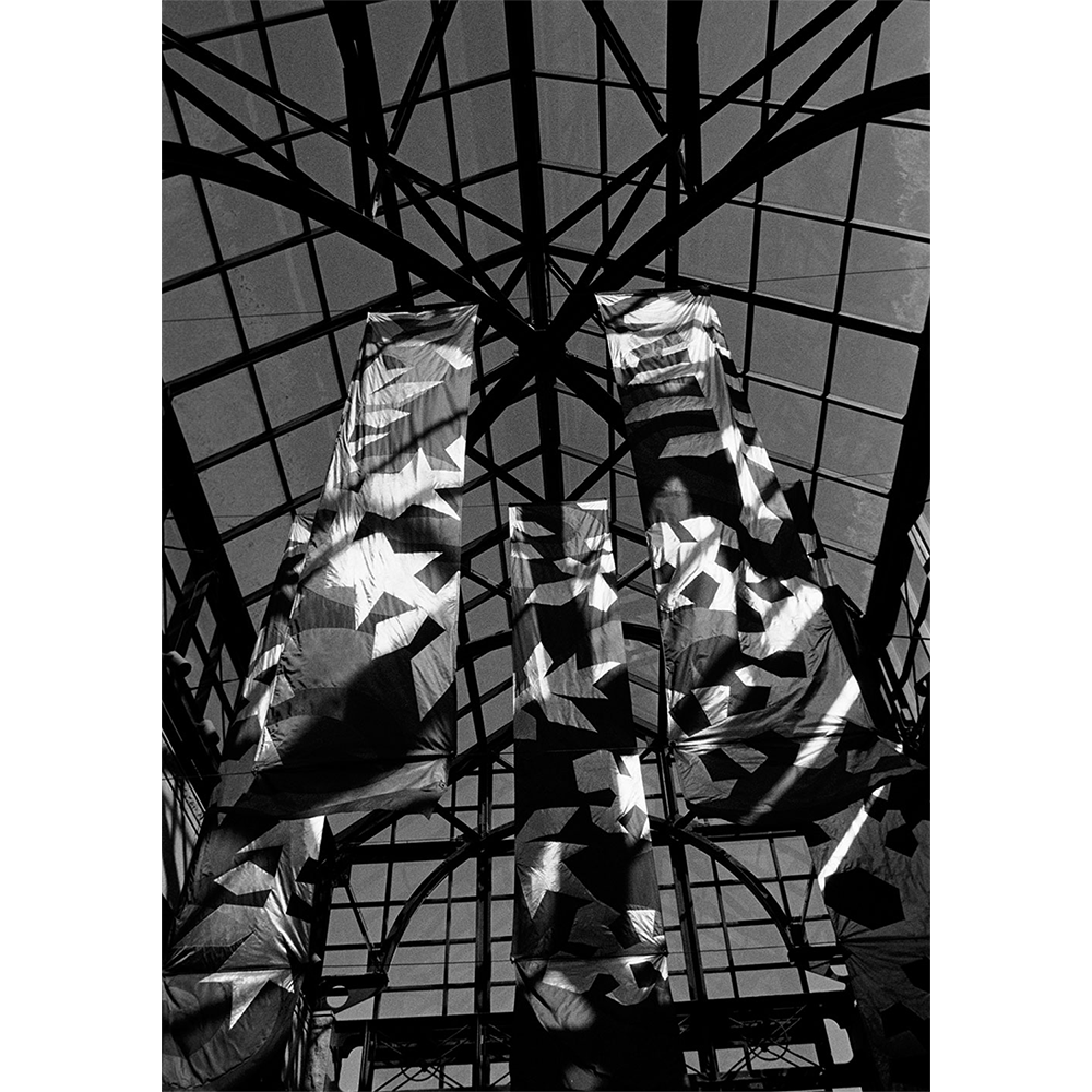 Silver gelatin photograph — star-patterned banners beneath a vaulted glass-and-steel atrium ceiling, hand-printed on vintage Brovira paper.