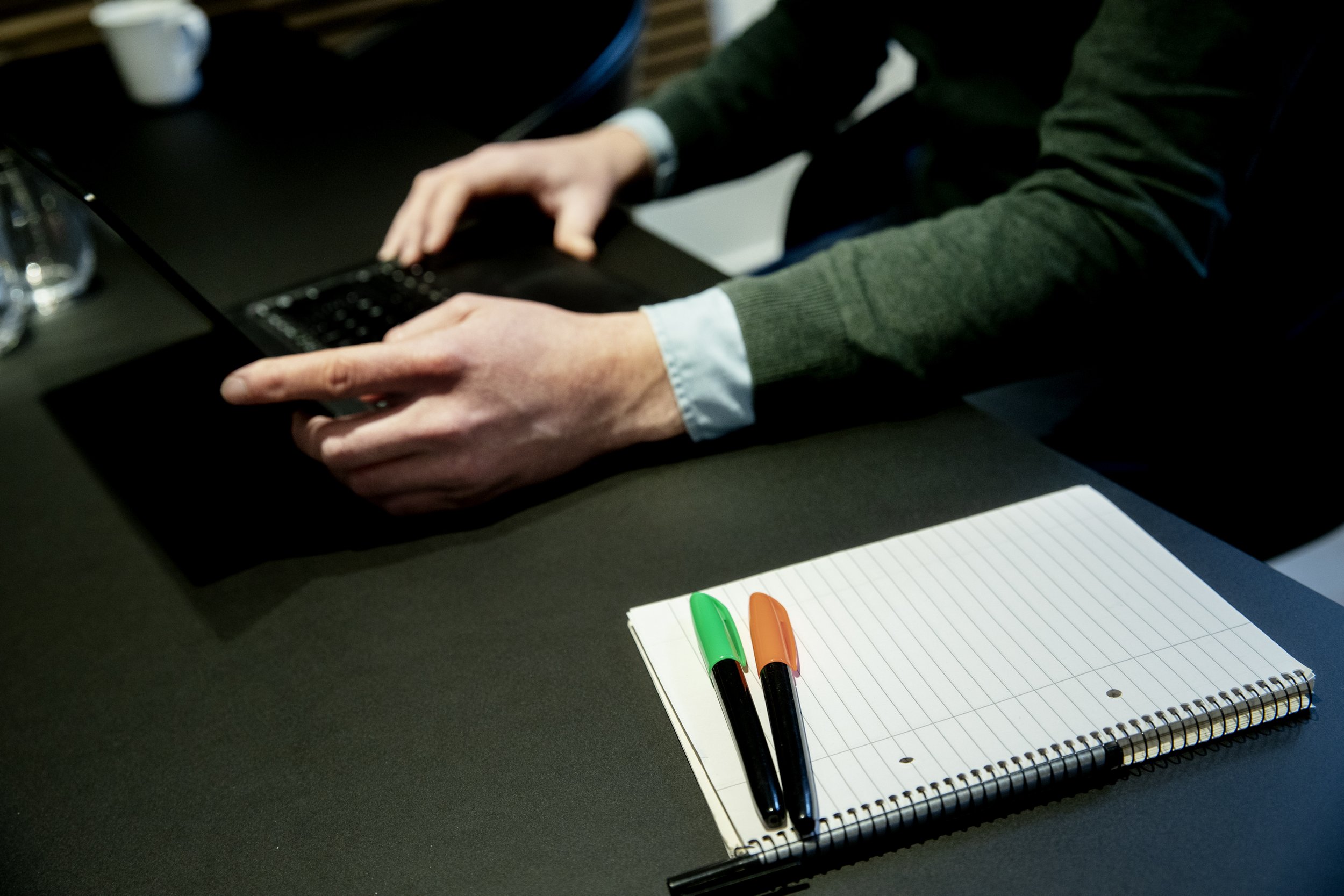 Person using a laptop with a notebook and two pens on the table.