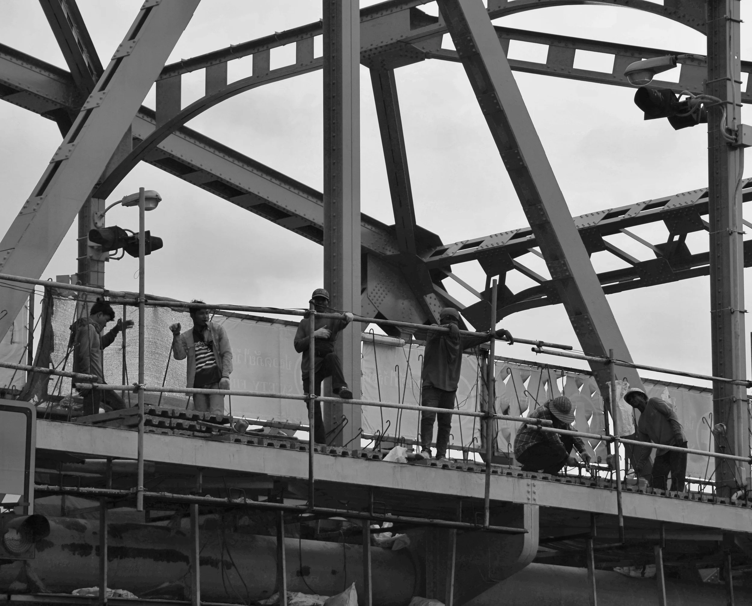 A group of construction workers on a platform working on a large steel structure, with some wearing helmets, under a cloudy sky.