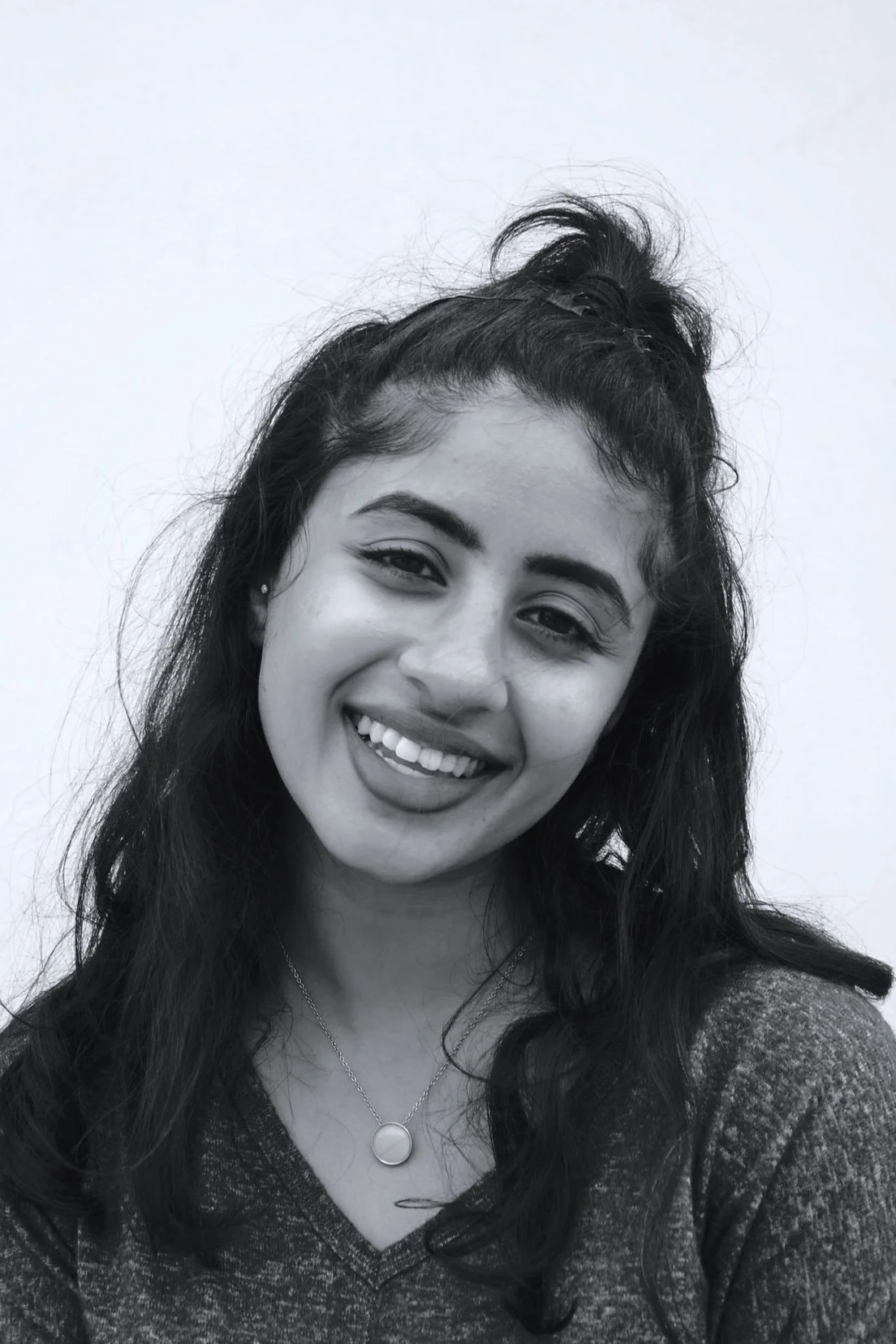 Black and white photo of a woman smiling with slightly tilted head, wearing a necklace and a textured top, with her hair up in a top knot and loose hair around her shoulders.
