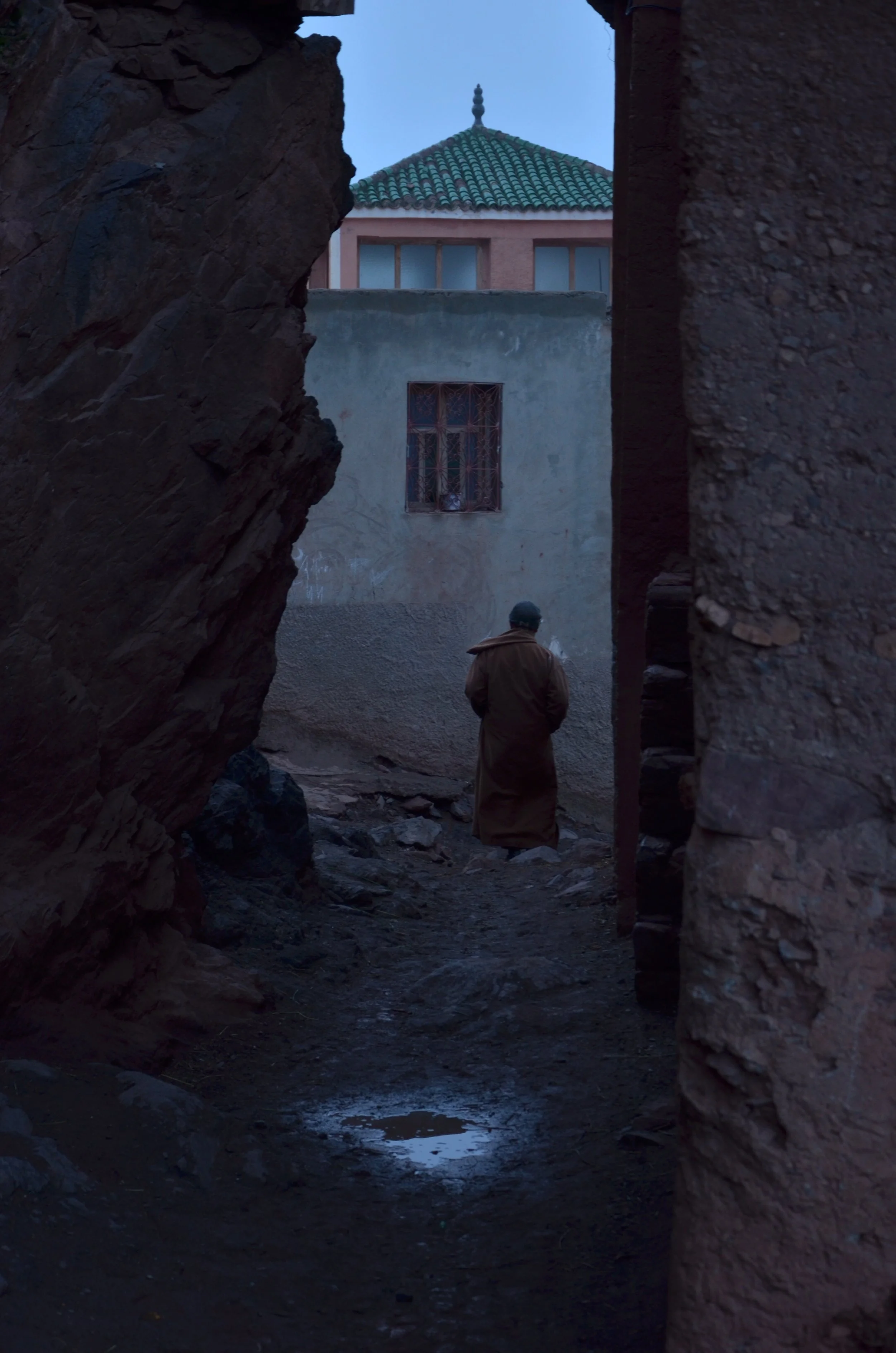 A person wearing a long coat and hooded hat walking through a narrow, rocky alleyway at dusk, with old buildings on either side and one building with a domed roof visible in the background.