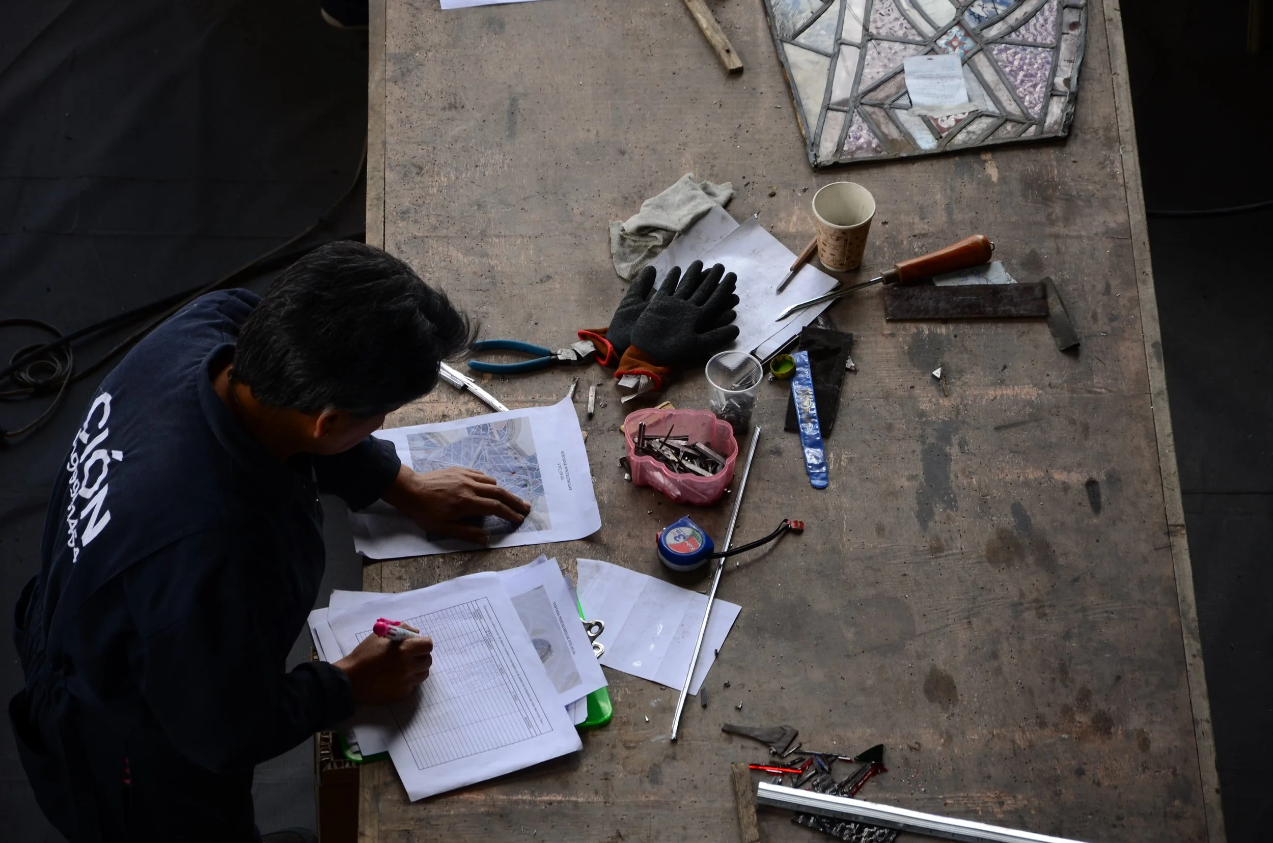 A person working at a wooden table, organizing and working on engineering or construction plans with various tools and materials scattered on the table.