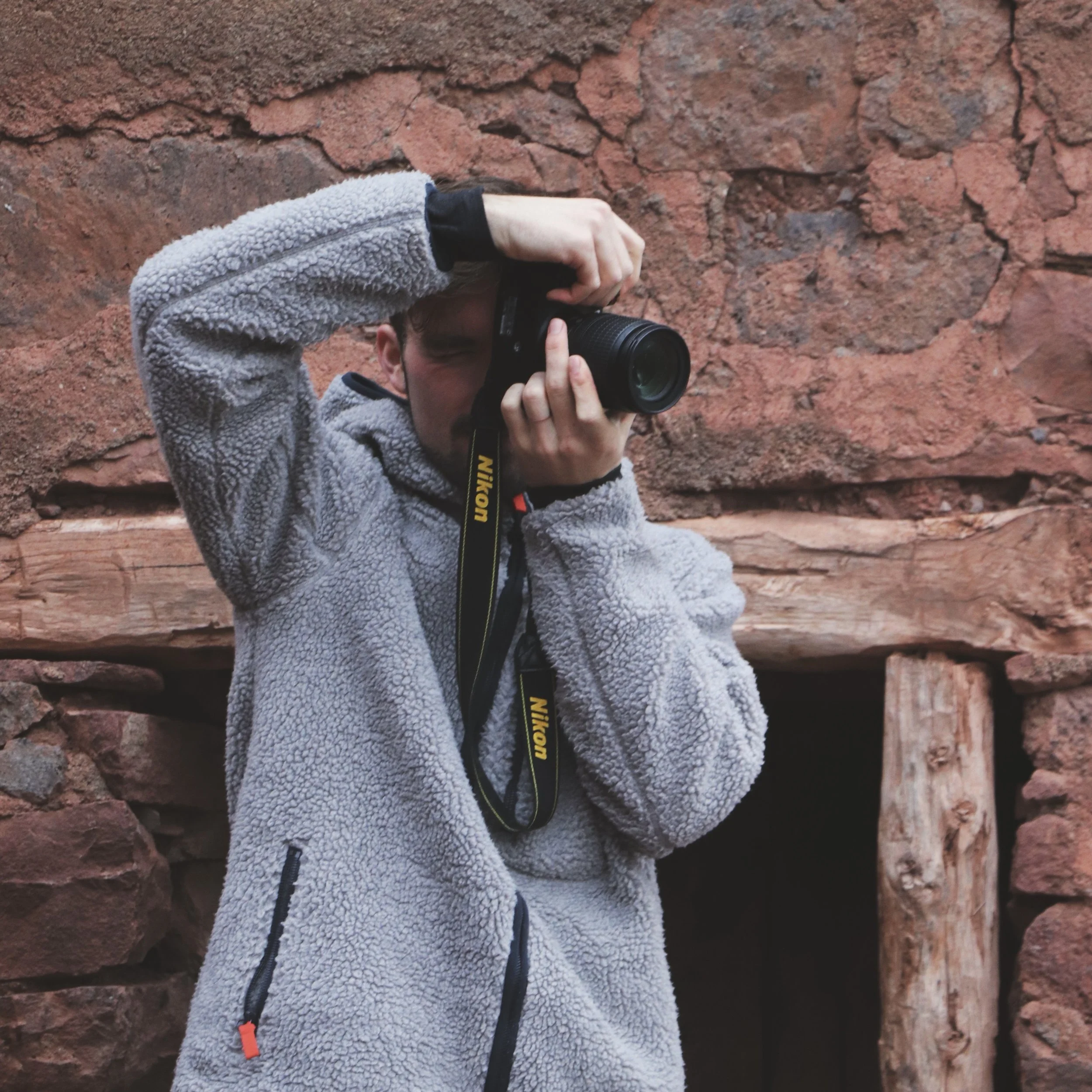 Person wearing a gray fleece jacket taking a photograph against a red rock background with a Nikon camera.