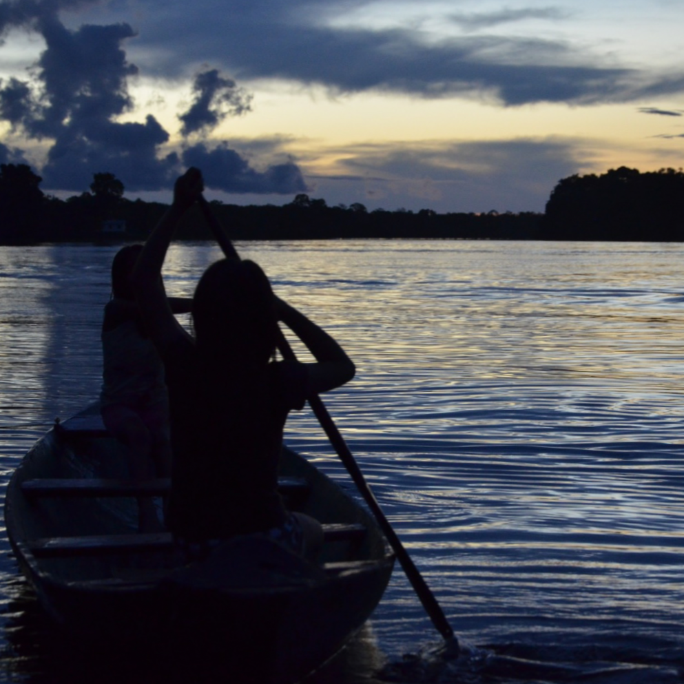 Silhouettes of two children paddling a small boat on a river during sunset with dark clouds in the sky.