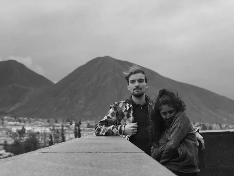 A black-and-white photo of a man and woman sitting outdoors with a mountain in the background, the woman is hugging her legs and leaning into the man.