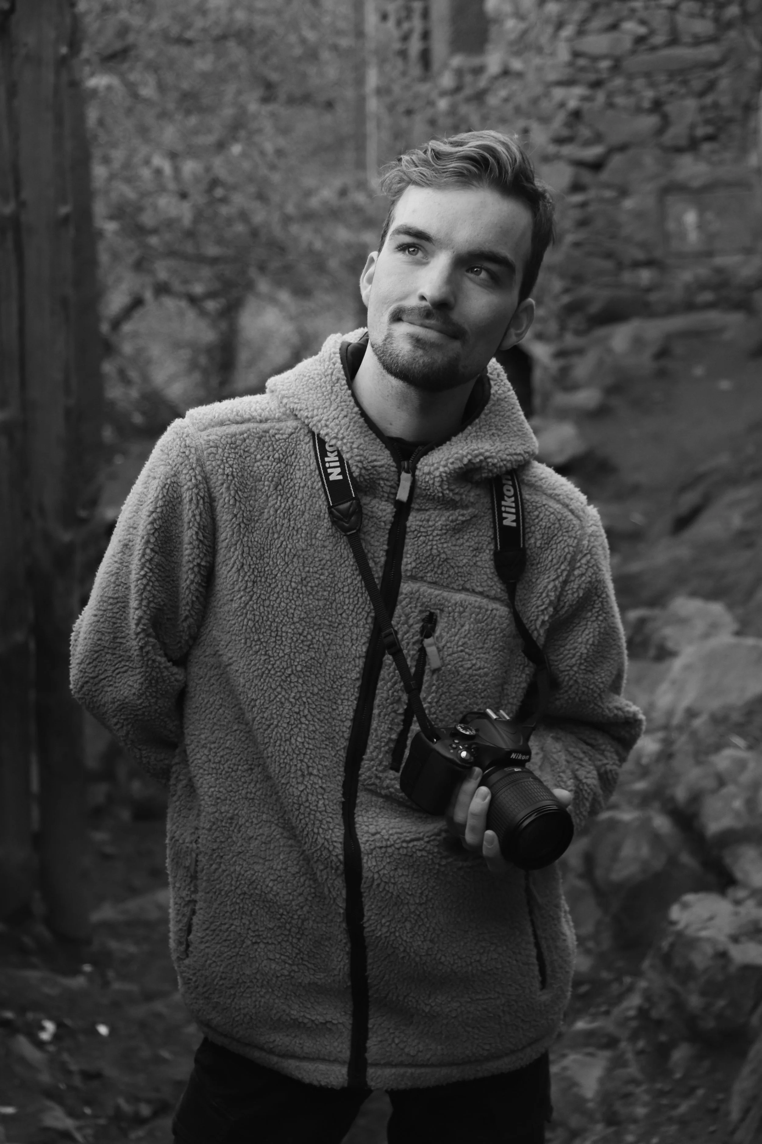 A young man standing outdoors on a trail, holding a camera, wearing a fleece jacket, and looking to the side in a thoughtful pose.
