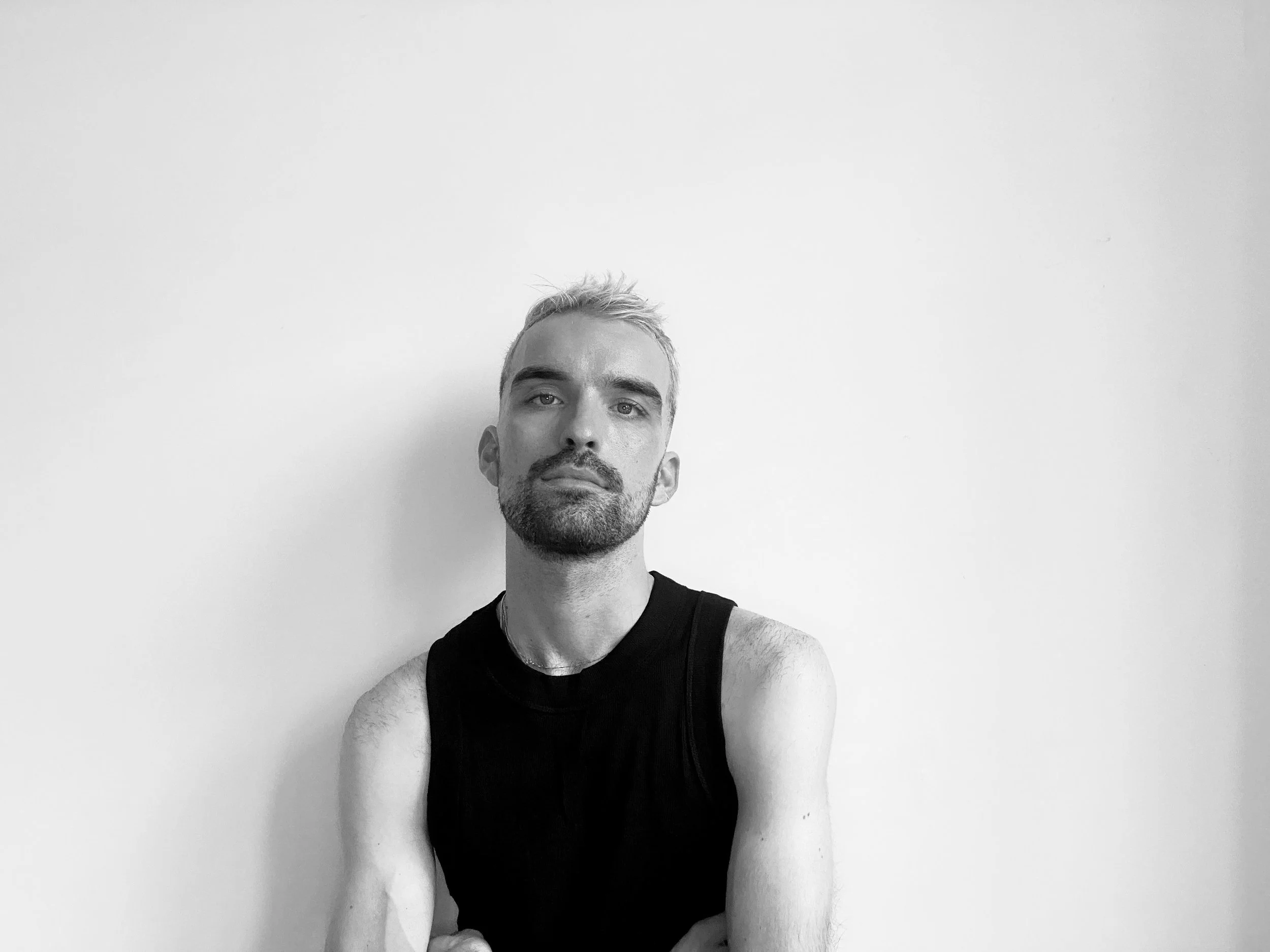 Black and white photograph of a young man with short, light-colored hair and a beard, wearing a sleeveless black shirt, standing against a plain light wall.