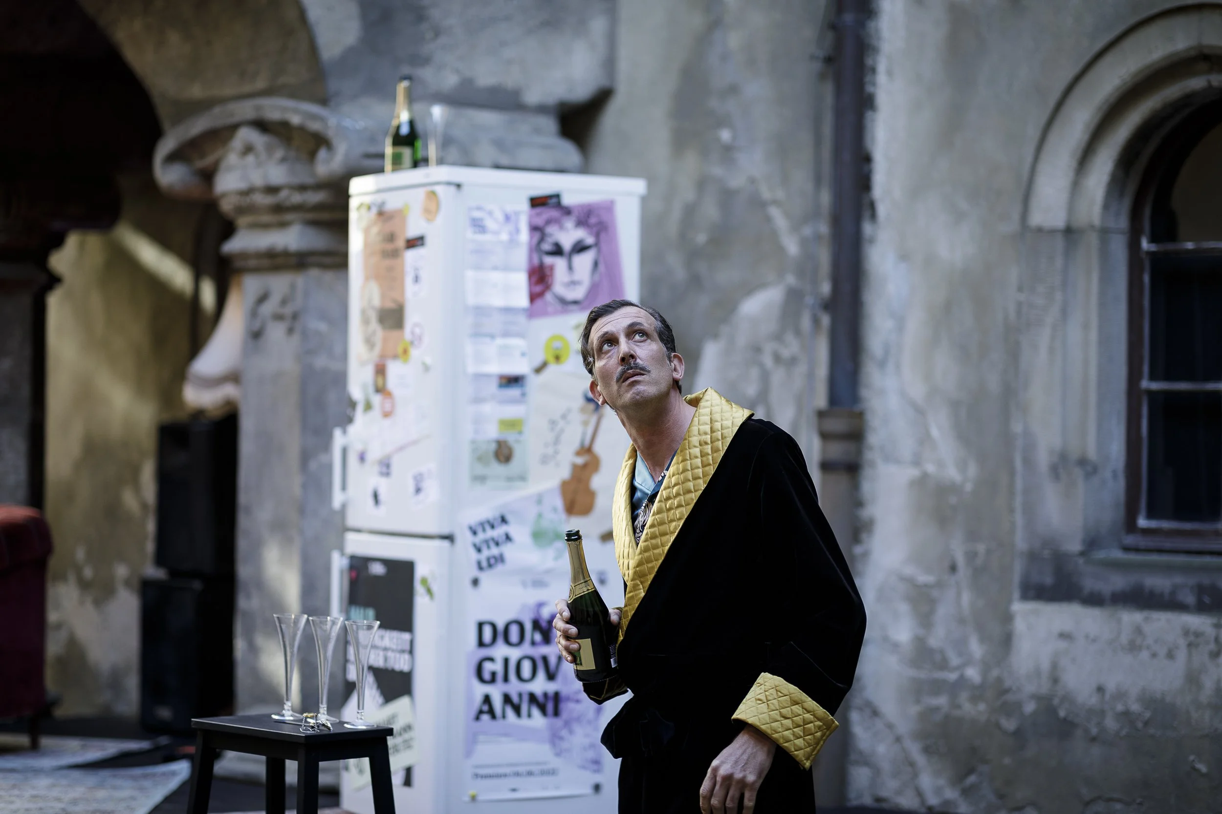 Ein Mann in einem Schwarz-Gold-Pullover hält eine Flasche Sekt und schaut nach oben. Im Hintergrund stehen drei leere Gläser auf einem kleinen Tisch, eine alte, graue Wand mit einem Fenster und eine Mülltonne.