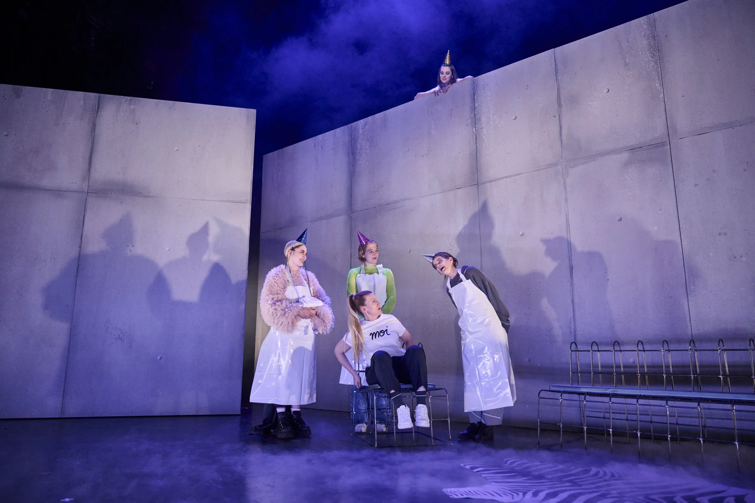 Szene aus einem Theaterstück mit fünf Frauen, von denen vier in weißen Schürzen und Partyhüten stehen, eine sitzt in einem Metallstuhl. Eine Frau oben auf einer Mauer schaut herab. Das Bühnenbild ist minimalistisch mit grauen Wänden. GRoße Schatten.