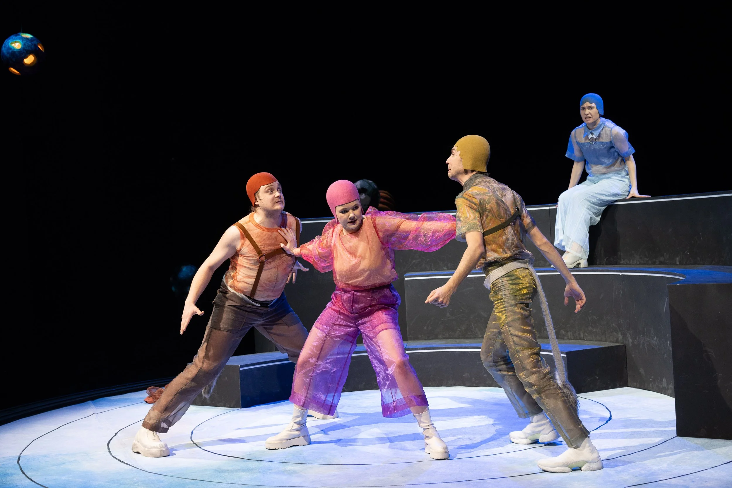 Theaterstück mit fünf Schauspielern in bunten Kostümen auf einer schwarz-weißen Bühne, eine Schauspielerin sitzt oben auf einem schwarzen Podest, vier sind unten in einer emotionalen Szene.