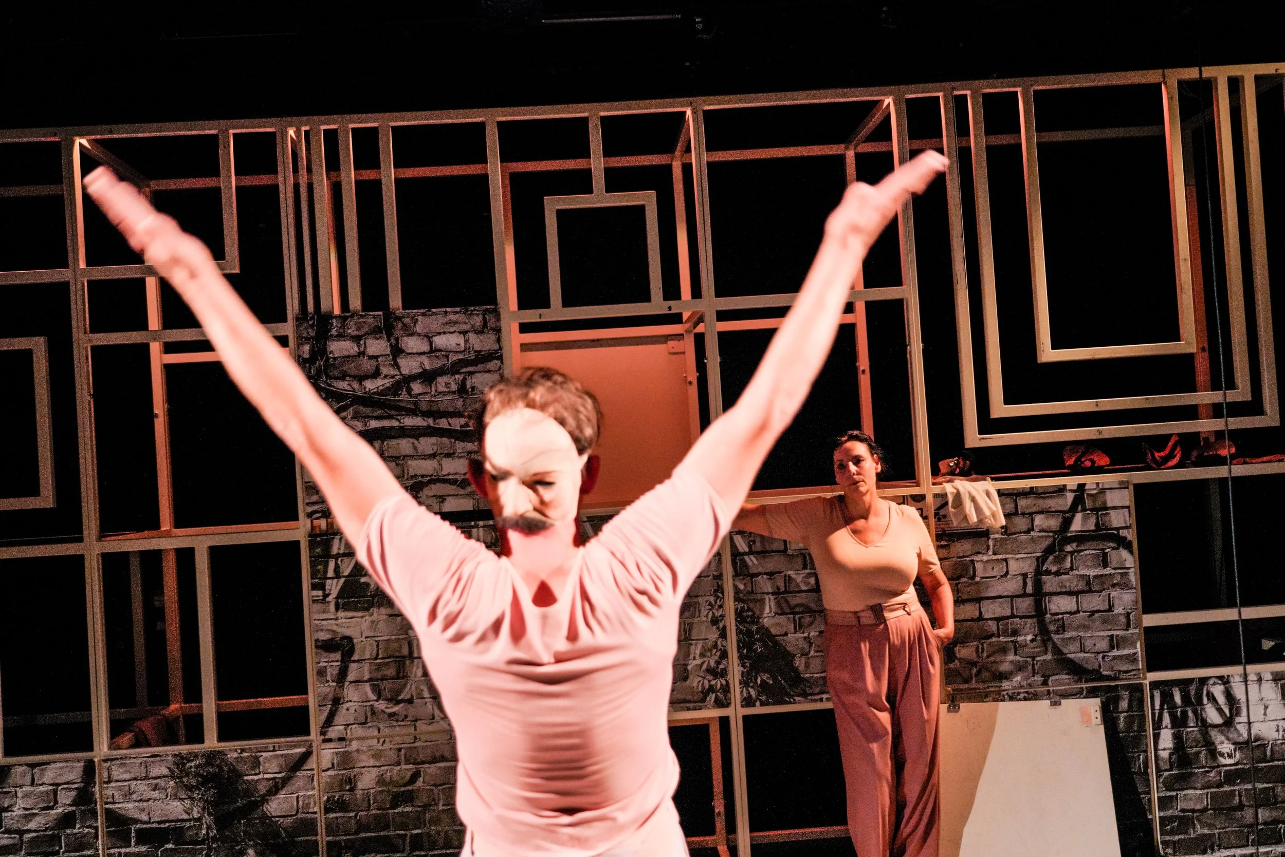 Zwei Frauen auf einer Bühne bei einer Theateraufführung. Die Frau im Vordergrund trägt ein rosa T-Shirt, eine falsche Nase und einen Schnurrbart, während die andere Frau im Hintergrund in einem beigen Top und rosa Hosen steht, vor einer Wand aus Holz