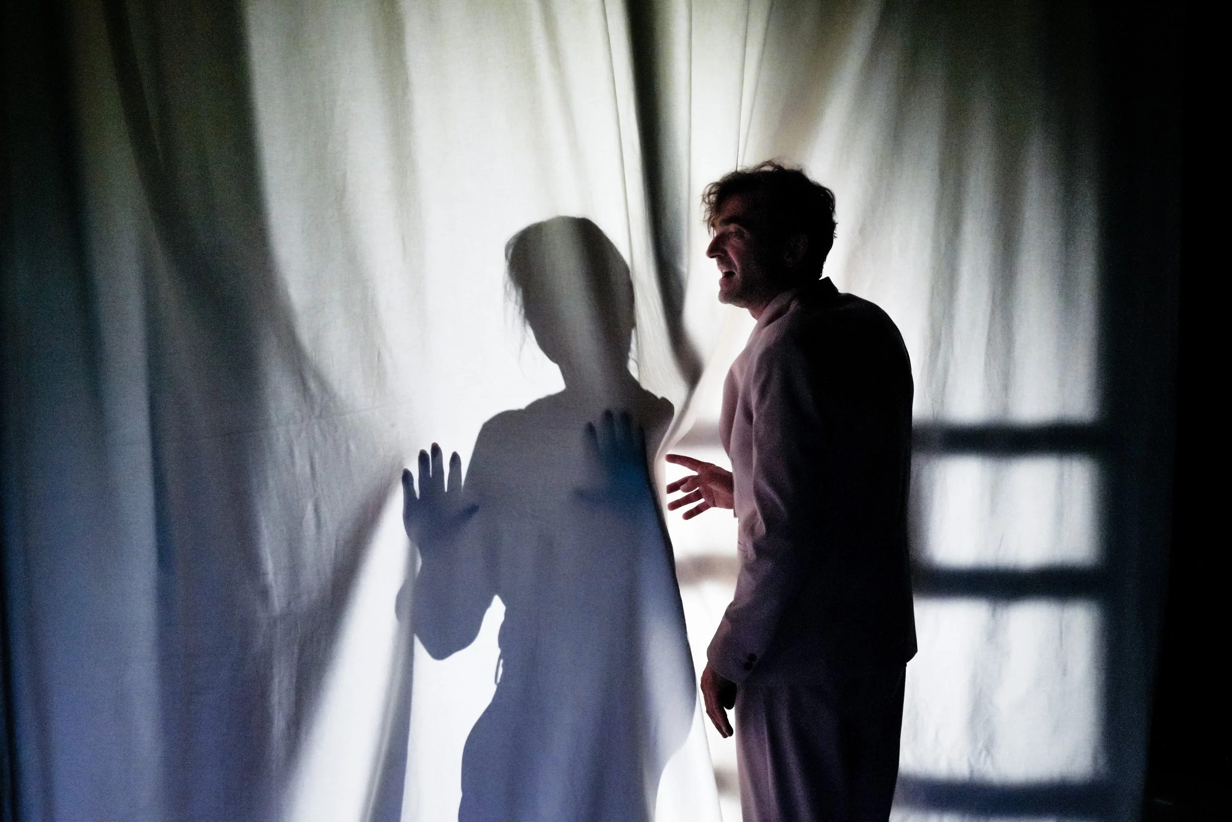 Ein Mann steht vor einem Vorhang und eine Frau stehen hinter einem Vorhang, der von Licht durchleuchtet wird, wodurch ihre Silhouetten sichtbar sind. Der Mann trägt einen Anzug und will den Schatten berühren. Mein Leben in Aspik in Regie von Drews