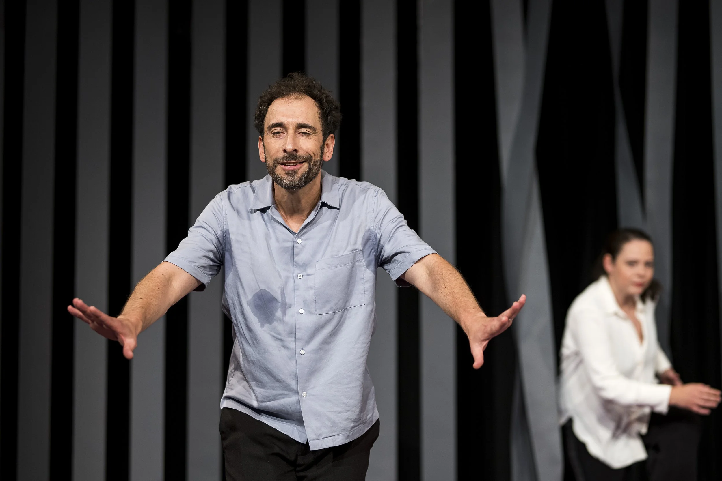 Zwei Menschen, ein Mann im Vordergrund und eine Frau im Hintergrund, auf einer Bühne mit dunklem, gestreiftem Hintergrund, die wahrscheinlich in einer Theaterszene oder einem Gespräch sind.