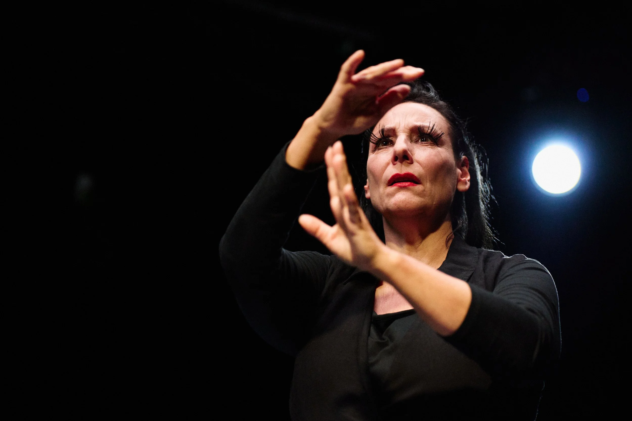 Eine Frau mit dunklem Haar und dunklem Kleid zeigt eine ausdrucksstarke Geste auf einer Bühne bei dunklem Hintergrund mit hellem Licht hinter ihr.
