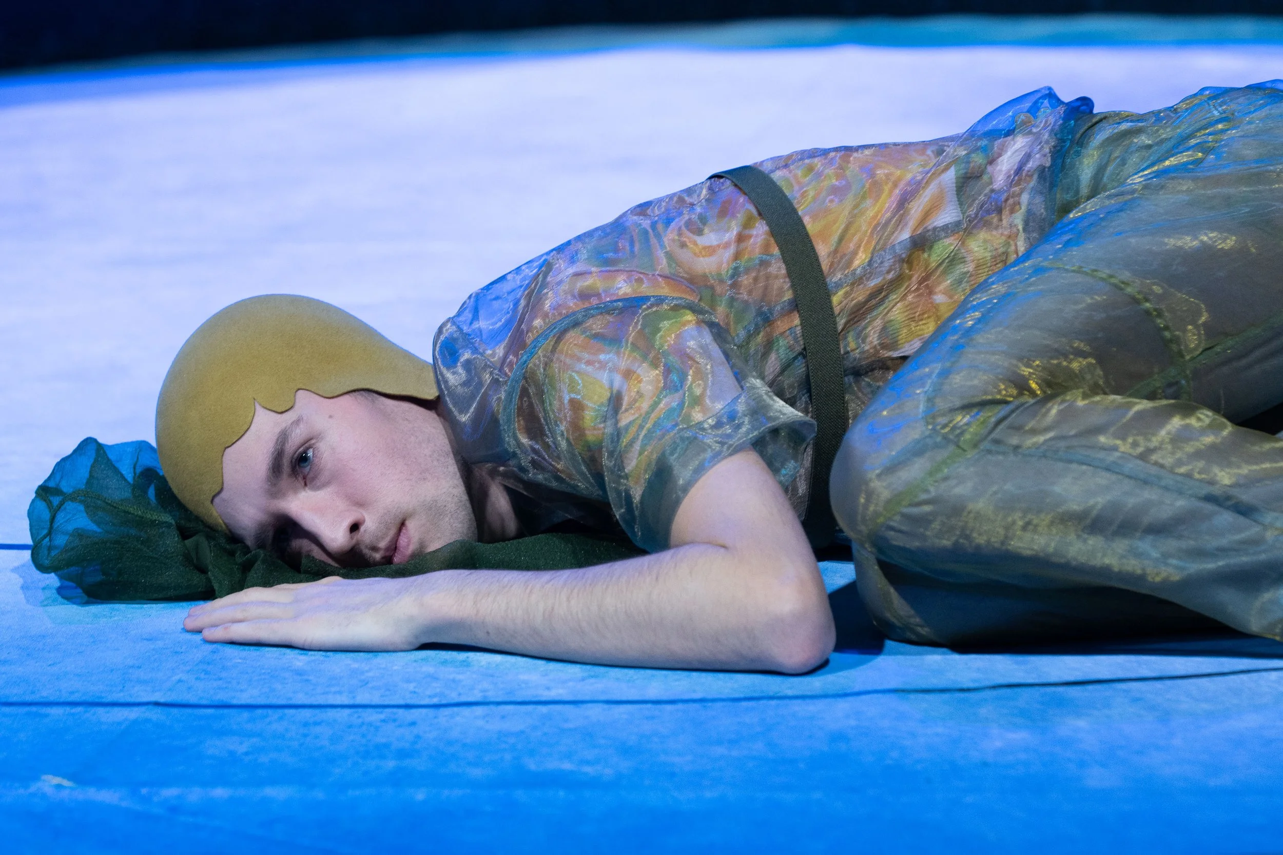 Junge Person mit gelbem Haarschmuck liegt auf dem Boden, trägt einen transparenten, bunten Overall, sieht nach dem Theaterstück aus, Szenenbild mit bläulichem Licht.