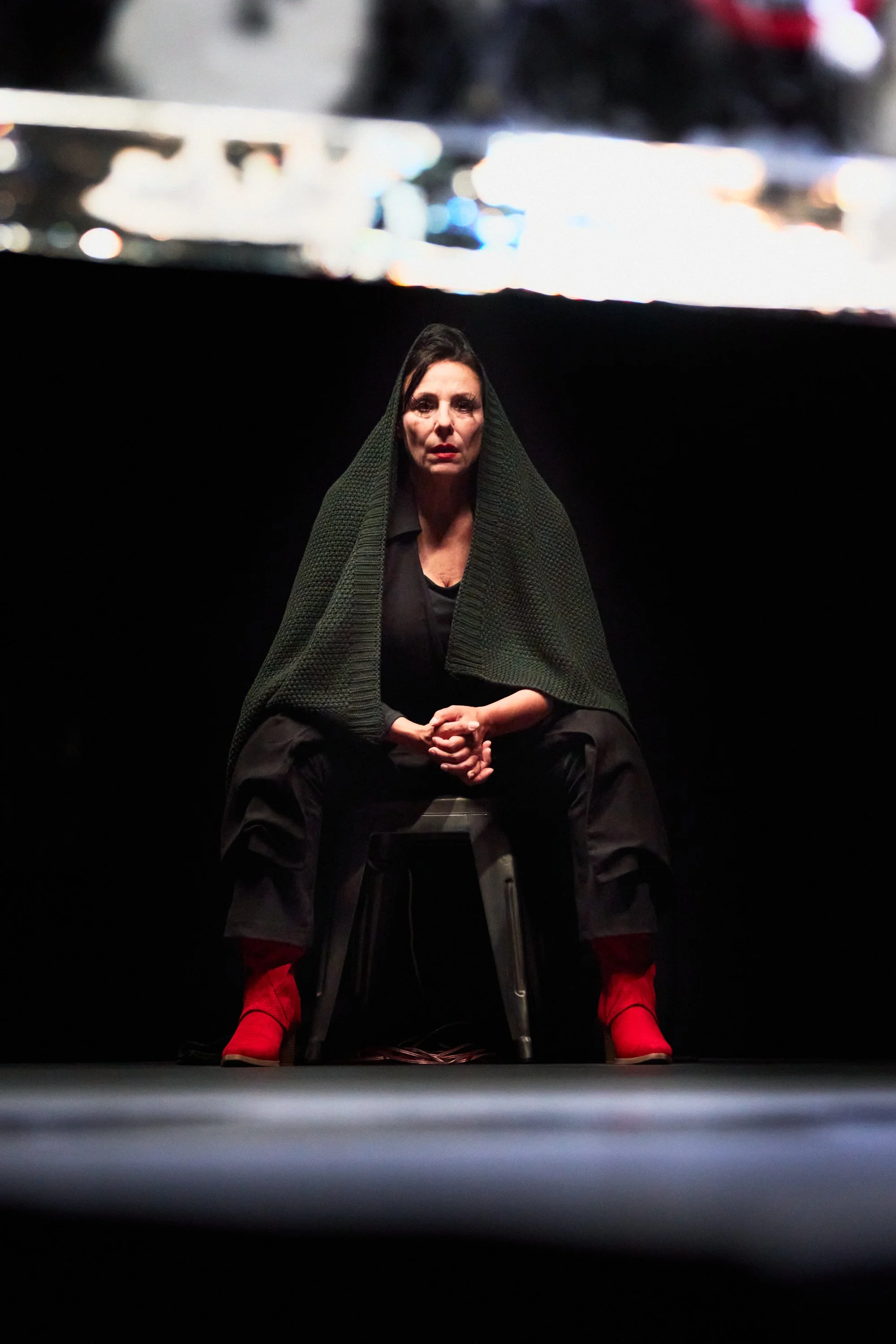 Frau sitzt auf einem Stuhl im Dunkeln, trägt eine dunkle Jacke, eine große grüne Decke über dem Kopf, rote Schuhe, im Hintergrund eine große, unscharfe Anzeige mit Lichtern.