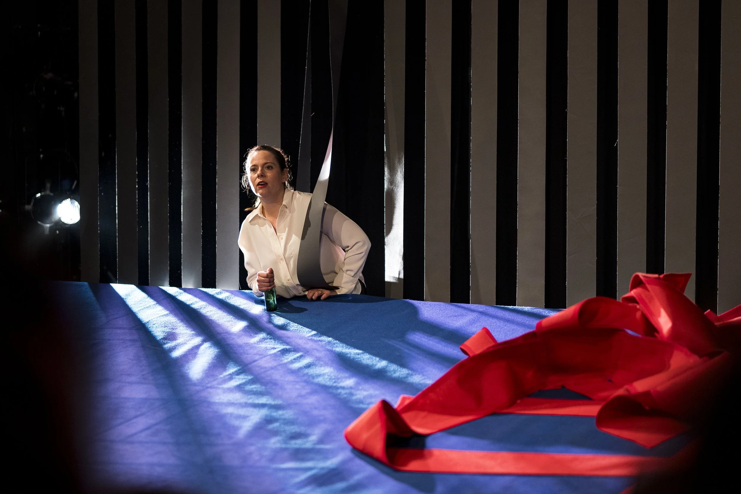 Eine Frau in weißem Hemd sitzt an einem langen Tisch, der mit blauer Tischtuch bedeckt ist. Es sind Streifen von Licht und Schatten auf dem Tisch. Im Vordergrund liegt ein zerknülltes rotes Band. Im Hintergrund sind schwarze vertikale Streifen und ei