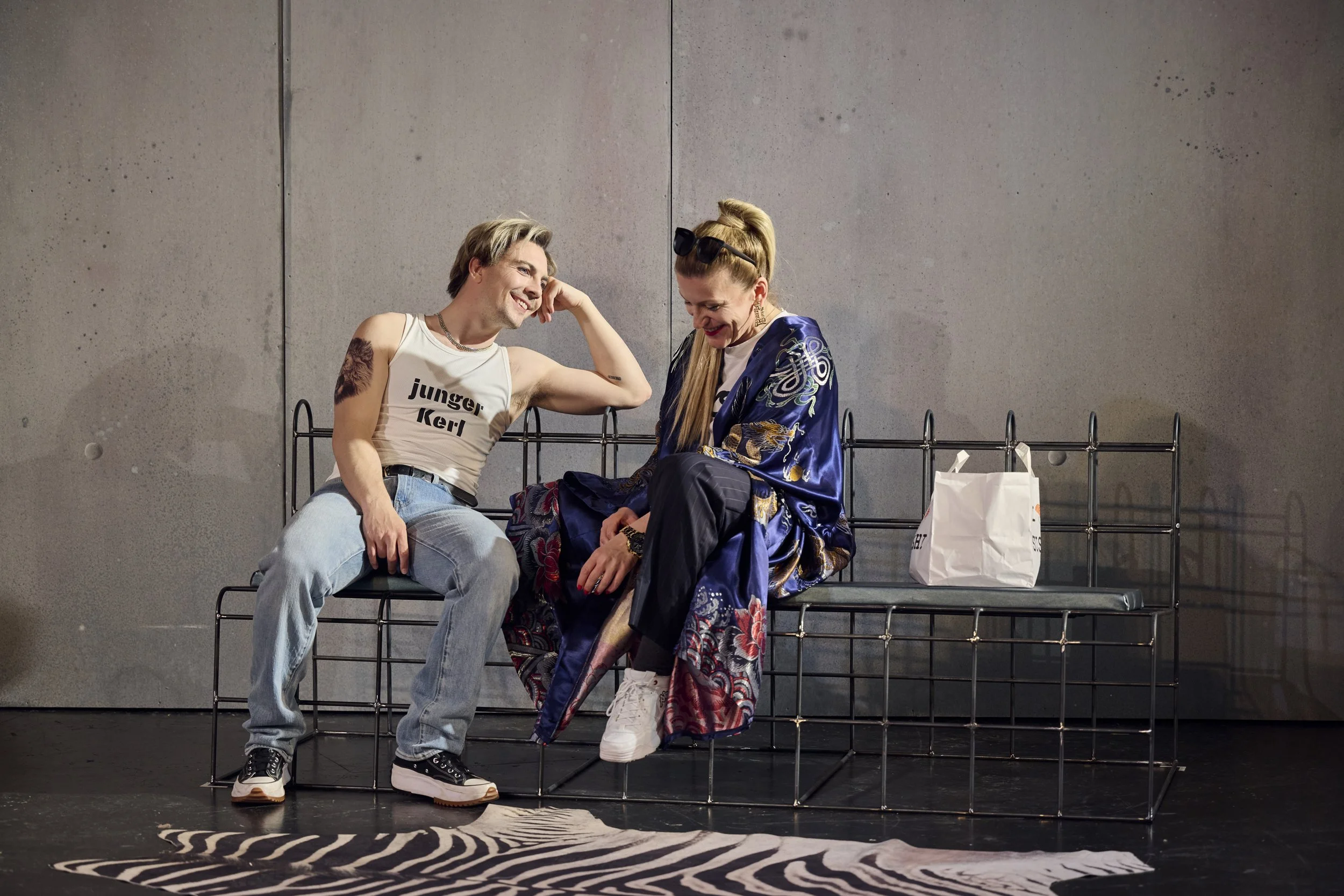 Zwei Menschen sitzen auf einer Bank gegen eine graue Wand, lachen und unterhalten sich. Die Person auf der linken Seite trägt ein weißes Tanktop und Jeans, die Person auf der rechten Seite trägt einen blauen, gemusterten Kimono. Es gibt eine weiße Ei