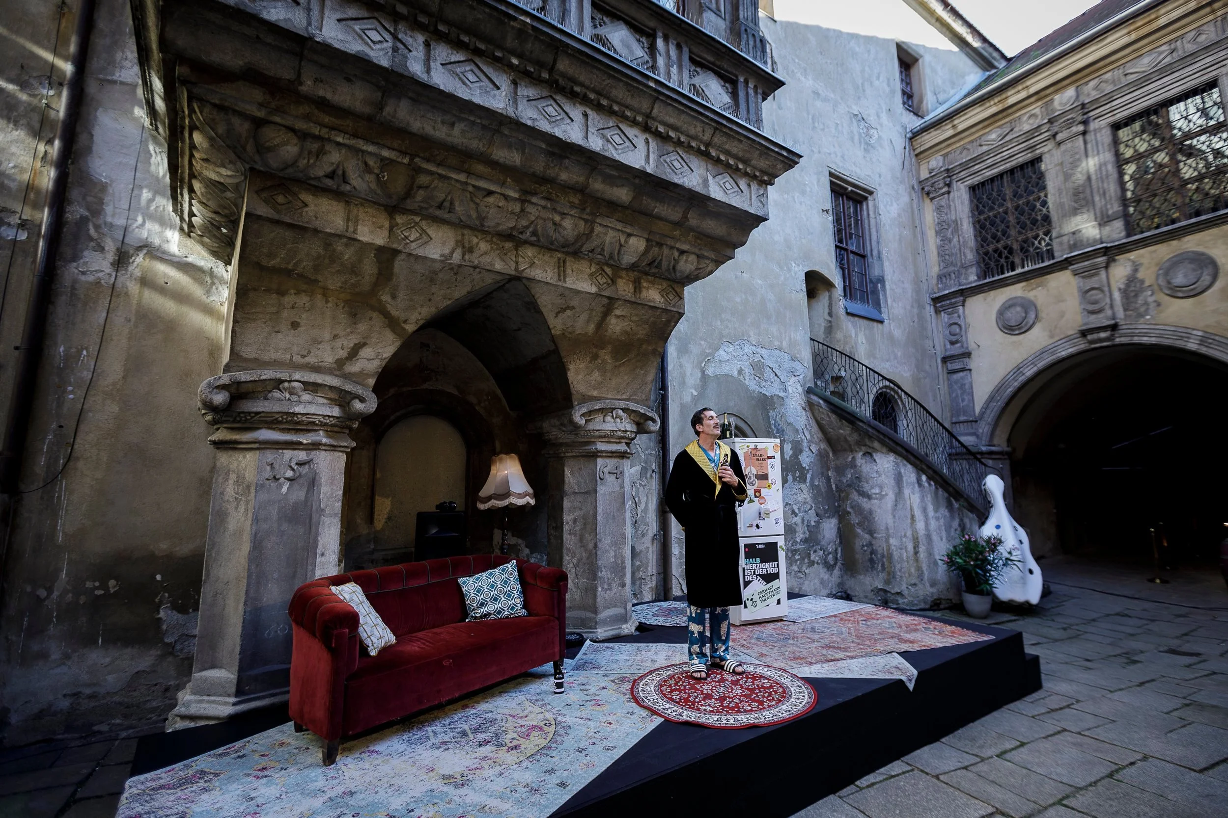Mensch auf einer kleinen Bühne in einem alten, verfallenen Innenhof, umgeben von Vintage-Möbeln und Dekorationen, mit einer Gitarre in der Hand.