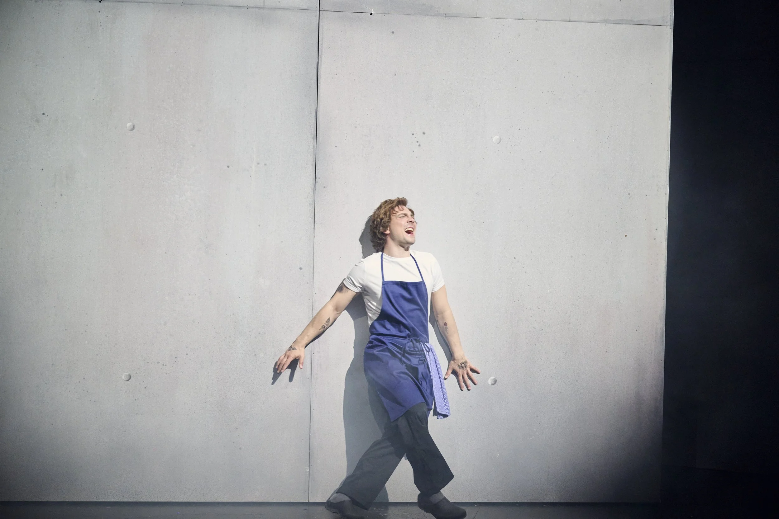 Ein junger Mann steht vor einer grauen Betonwand, trägt eine weiße T-Shirt und eine blaue Schürze, gestikuliert dramtaisch. Er sieht aus wie aus der Serie THE BEAR.