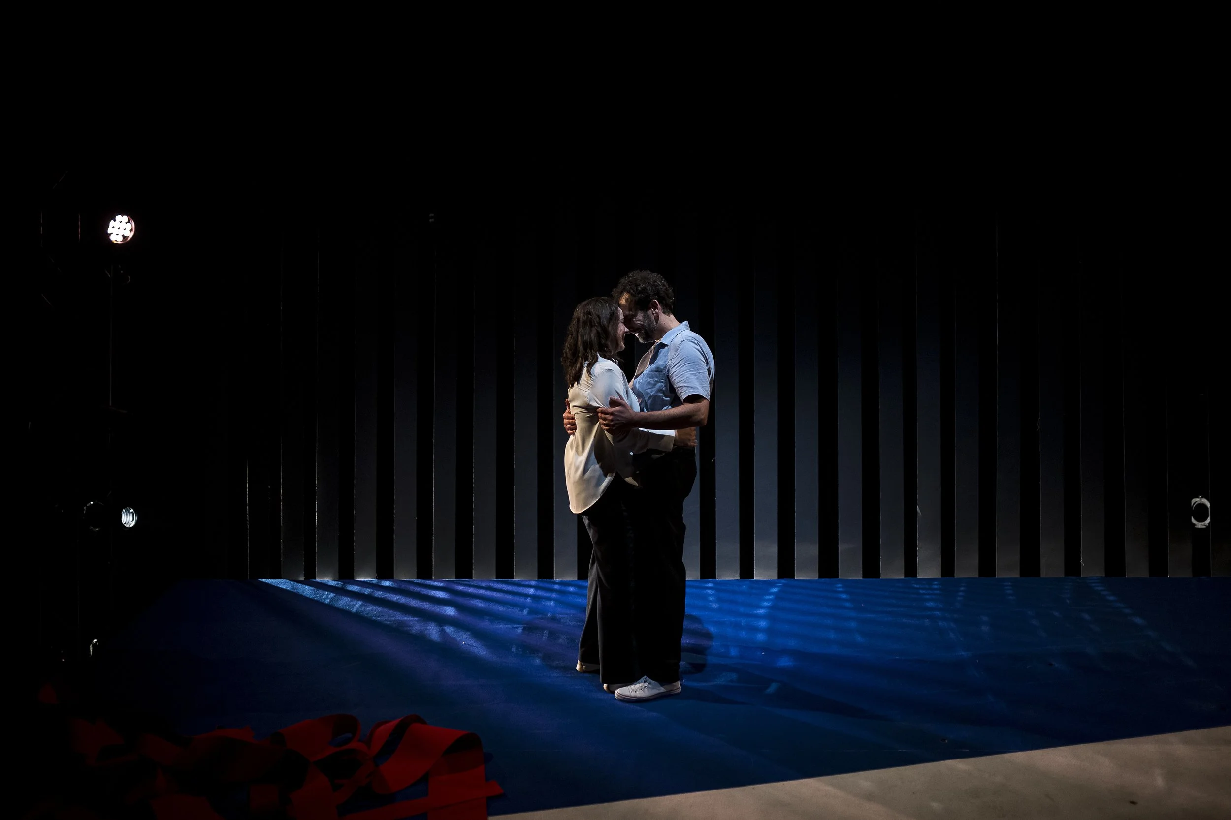 Zwei Menschen stehen sich auf einer dunklen Bühne nah gegenüber, in einer intimen Umarmung, mit einem schwarzen Hintergrund und seitlichem Licht