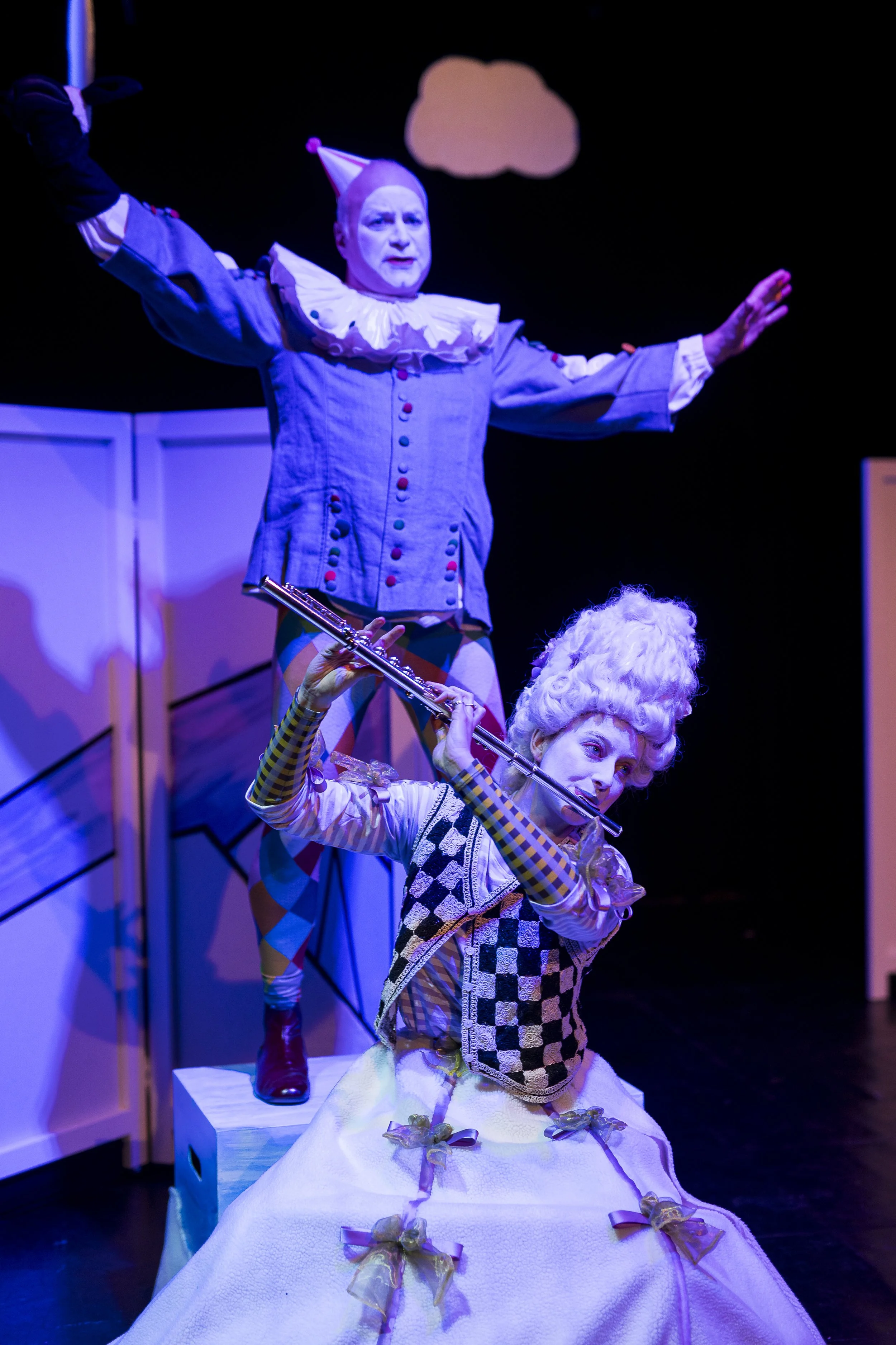 Zwei Clowns, einer mit Flöte und Kopfbedeckung, posieren auf einer Bühne mit bunter Kleidung und einem romantisch dekorierten Hintergrund bei einer Theatervorstellung.