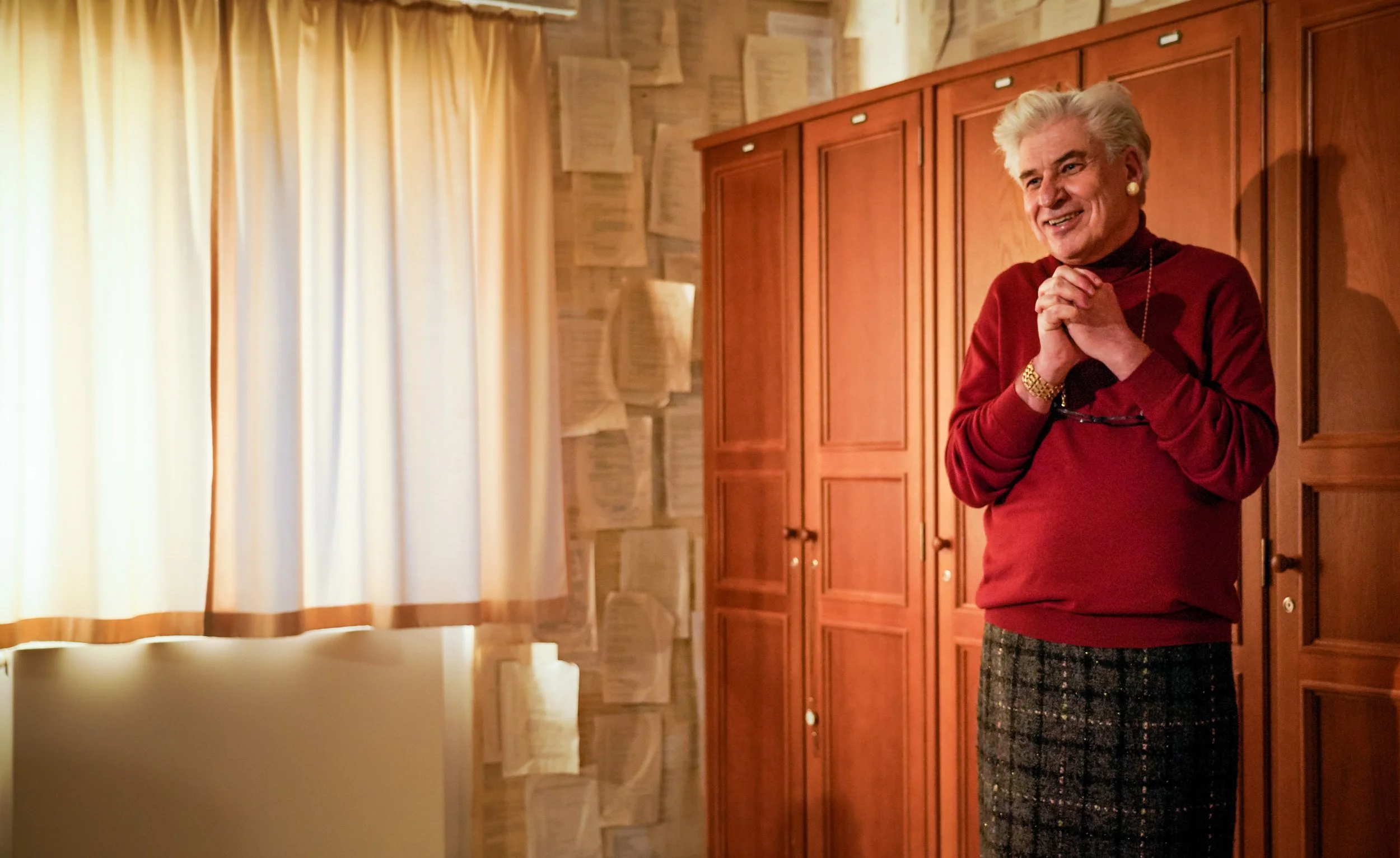 Ältere Frau mit weißen Haaren, rotem Pullover, schwarzer Hose und goldenen Accessoires steht vor einem Holzschrank in einem Raum mit beige Gardinen und Papier an der Wand, lächelt und sieht glücklich aus.