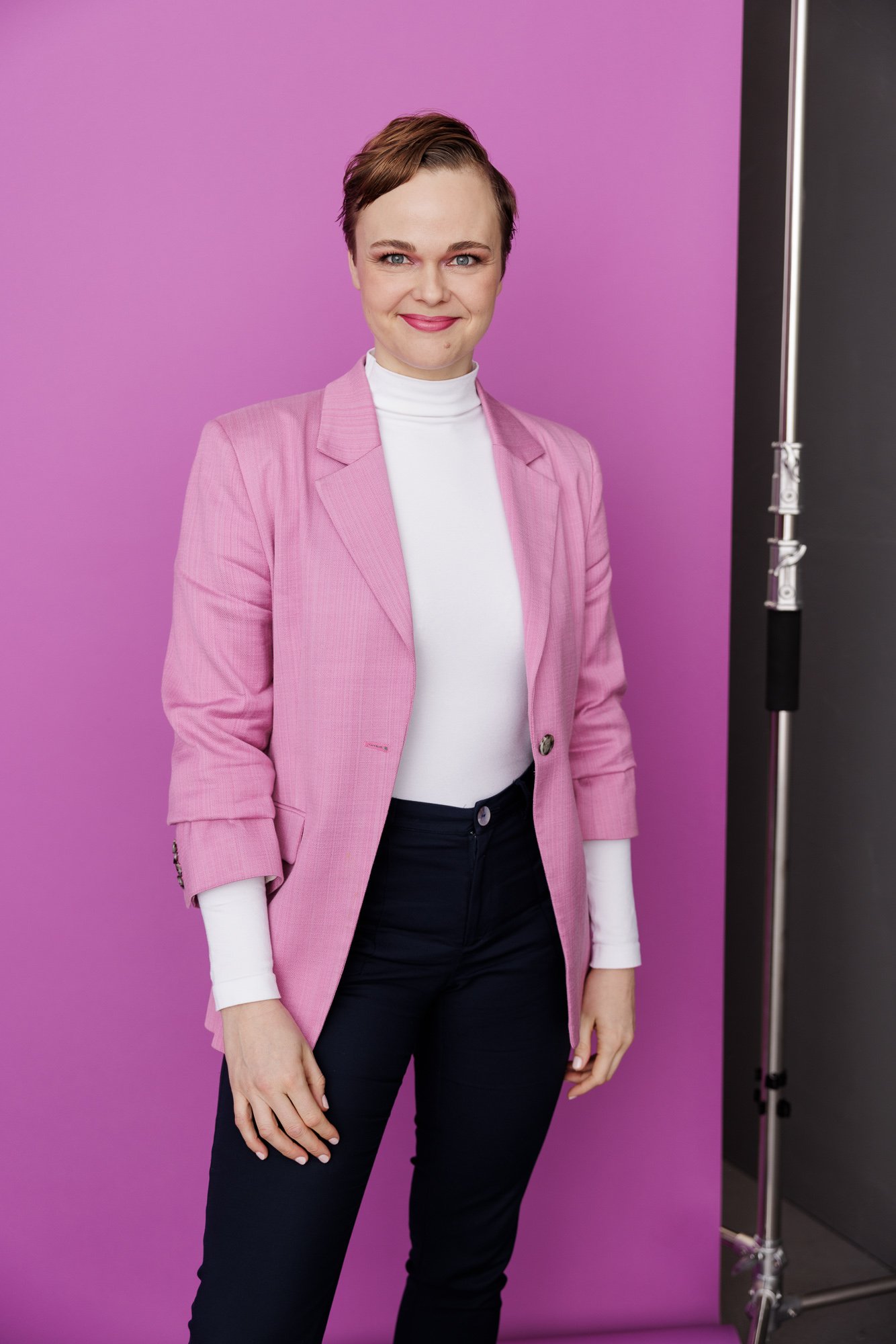 Junge Frau mit Kurzhaarfrisur trägt pinke Blazer, weißen Rollkragenpullover und dunkle Hosen vor pinkem Hintergrund. Die Regisserin Friederike Drews posiert wie eine Managerin.
