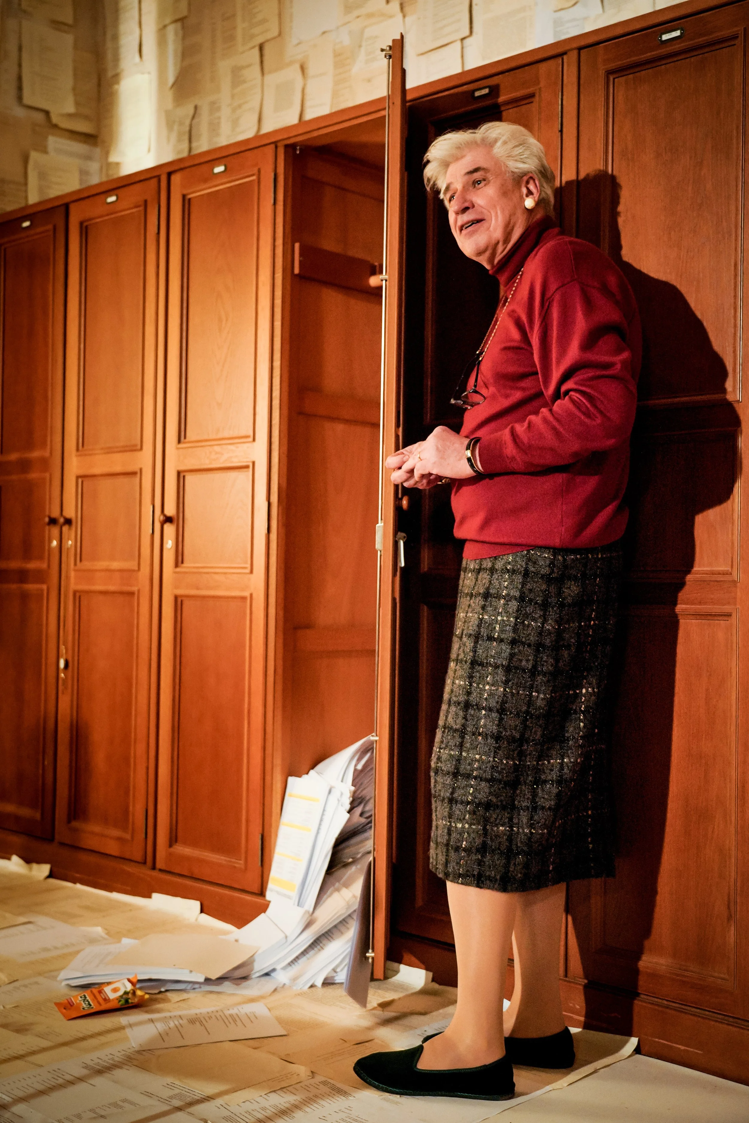Ältere Frau mit grauem Haar, rotem Pullover, kariertem Rock, schwarzen Schuhen, steht neben braunem Wandschrank, der offen ist, vor einem Boden voller Papierstapel, die Papier- und Zeitschriftenseiten enthalten.