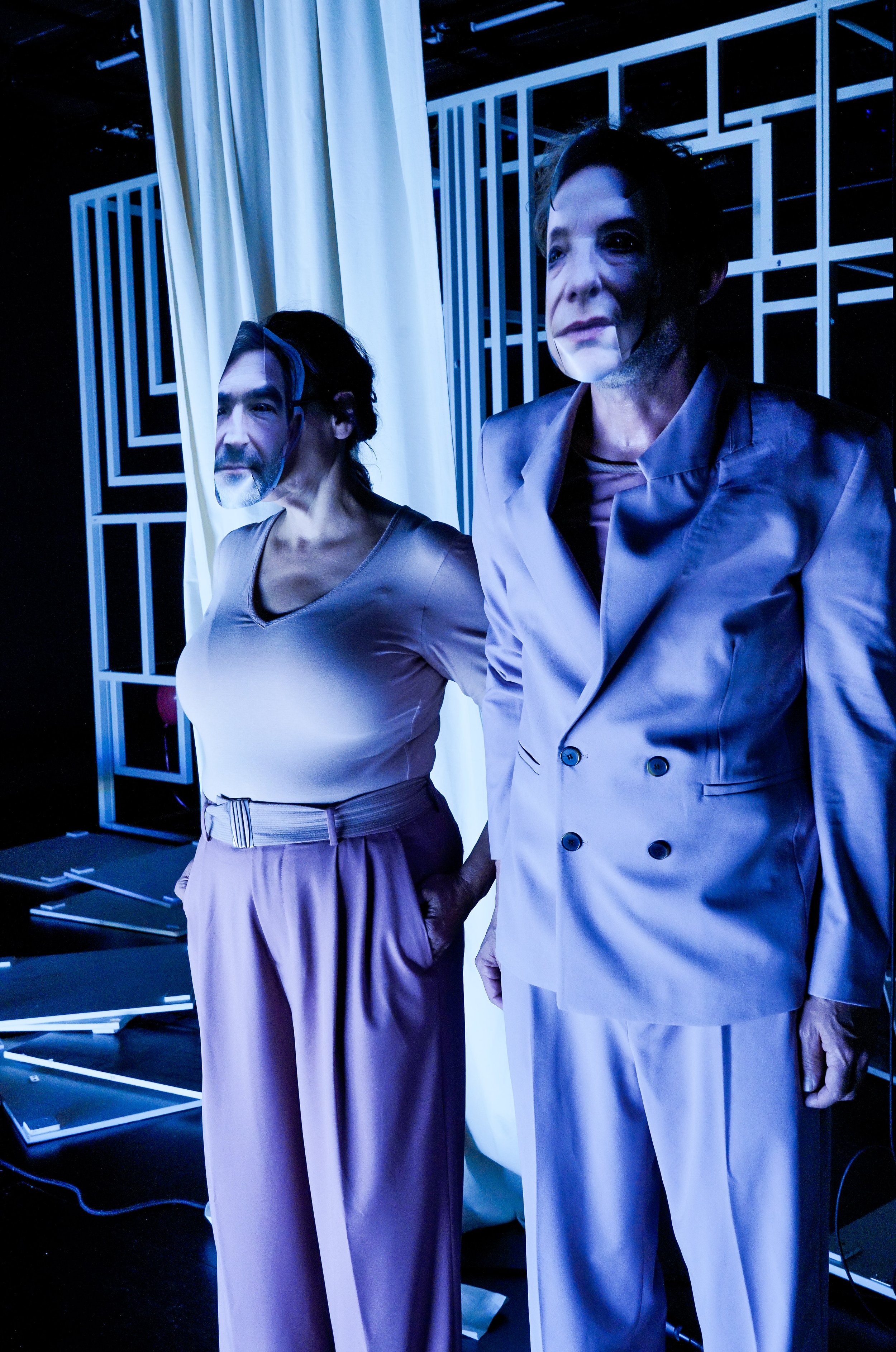 Zwei Personen in modernen Kleidung stehen nebeneinander vor einer geometrischen Kulisse; der eine trägt eine hellgraue Anzugjacke, der andere ein helles Top mit langen Hosen.