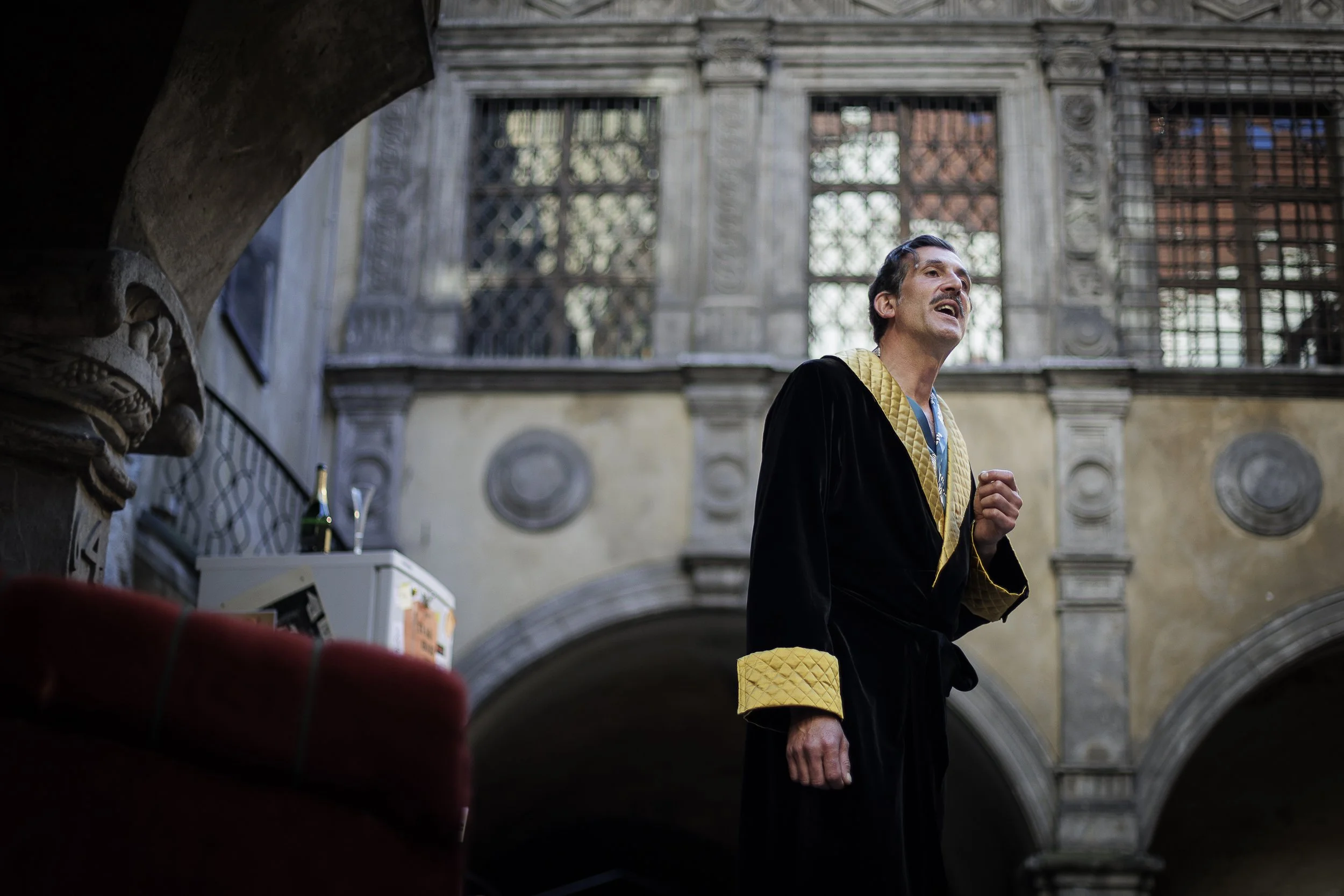 Ein Mann in einem schwarzen Bademantel mit goldenen Innenfutter singt in einem historischen Innenhof, umgeben von alten steinernen Gebäuden und Gittern an den Fenstern.