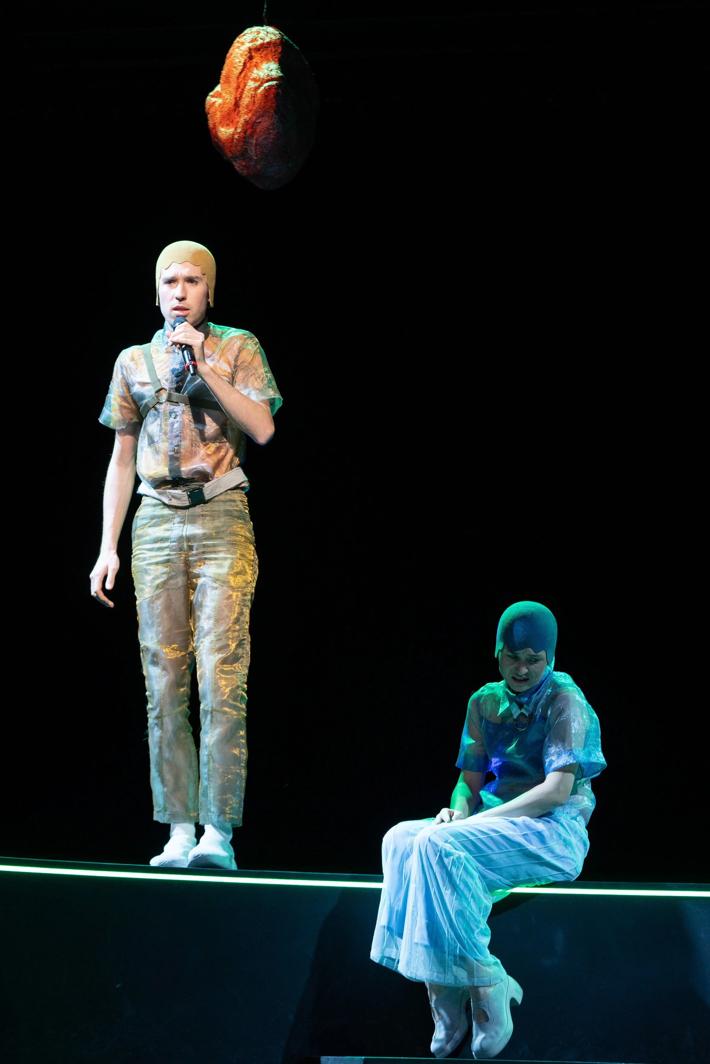 Zwei Schauspieler in bunten Kostümen auf einer Bühne mit dunklem Hintergrund, einer steht und spricht in ein Mikrofon, die andere sitzt und schaut nach unten, über ihnen schwebt ein großer, roter, unregelmäßiger Felsen.
