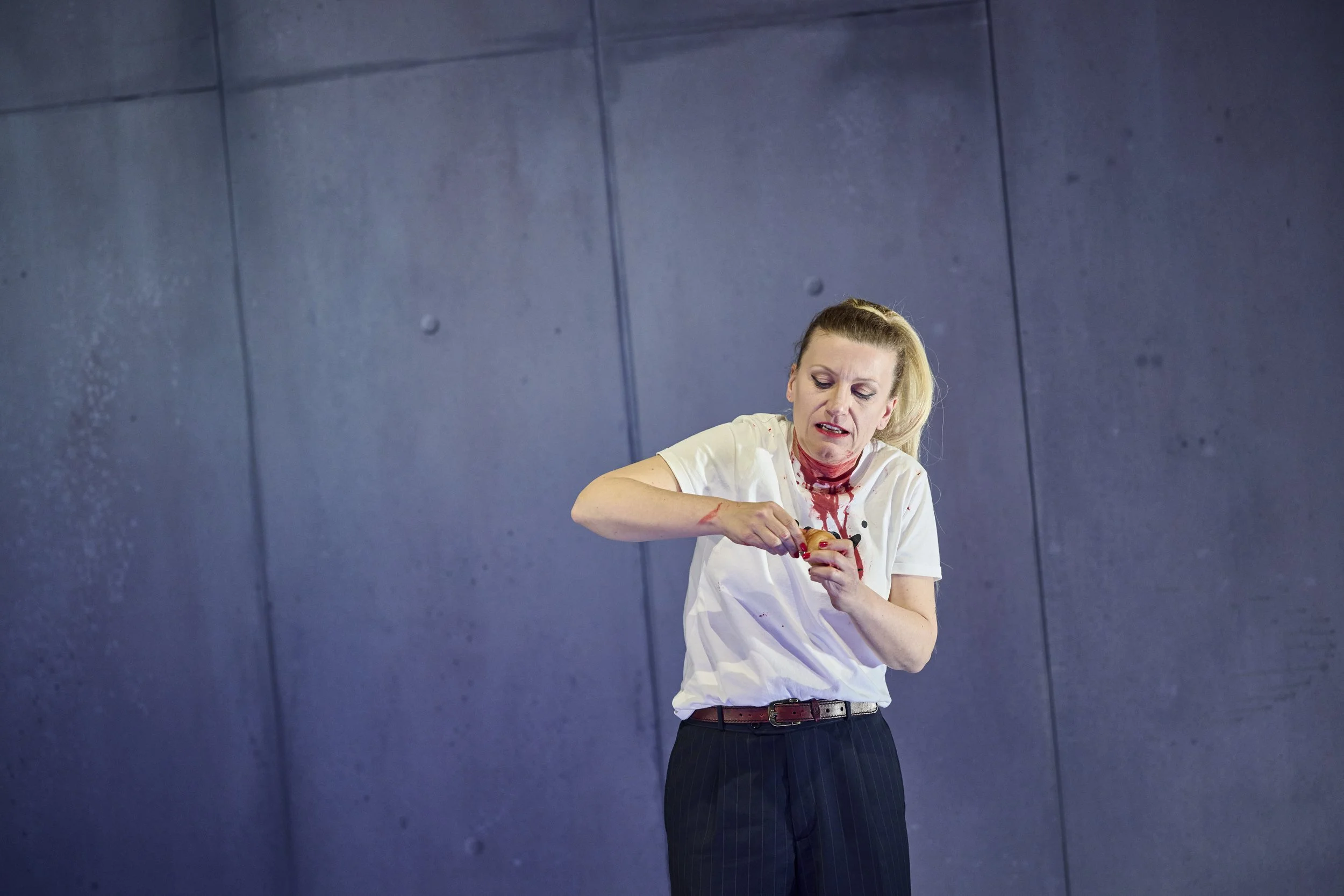 Theaterdarstellerin mit blutigem Hemd und Gesichtsausdruck des Schmerzes steht vor grauer Wand und schält eine Zwiebel. Landestheater Tübingen. Regie Friederike Drews Bühne Ev Benzing.