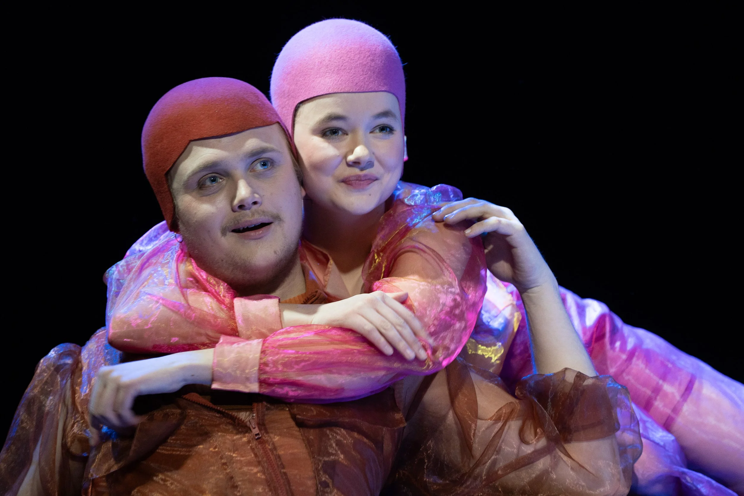 Zwei Schauspieler in bunten Posen, einer umarmt den anderen. Beide tragen farbige, glitzernde Kostüme und Kopfbedeckungen, die an Haarfarben erinnern.