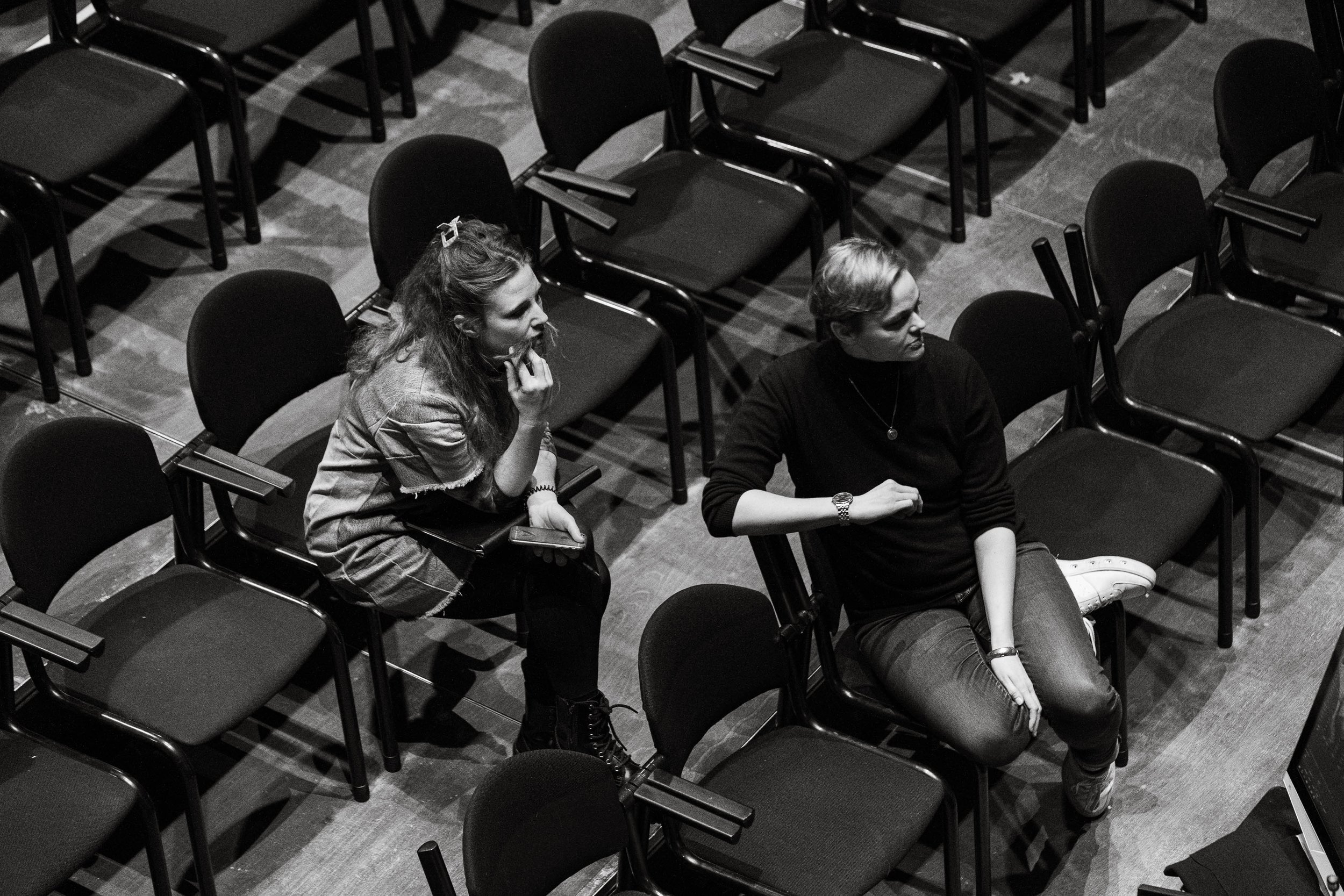 Zwei weibliche Personen sitzen in einem leeren Saal auf einzelnen schwarzen Stühlen, seitlich betrachtet, in Schwarz-Weiß