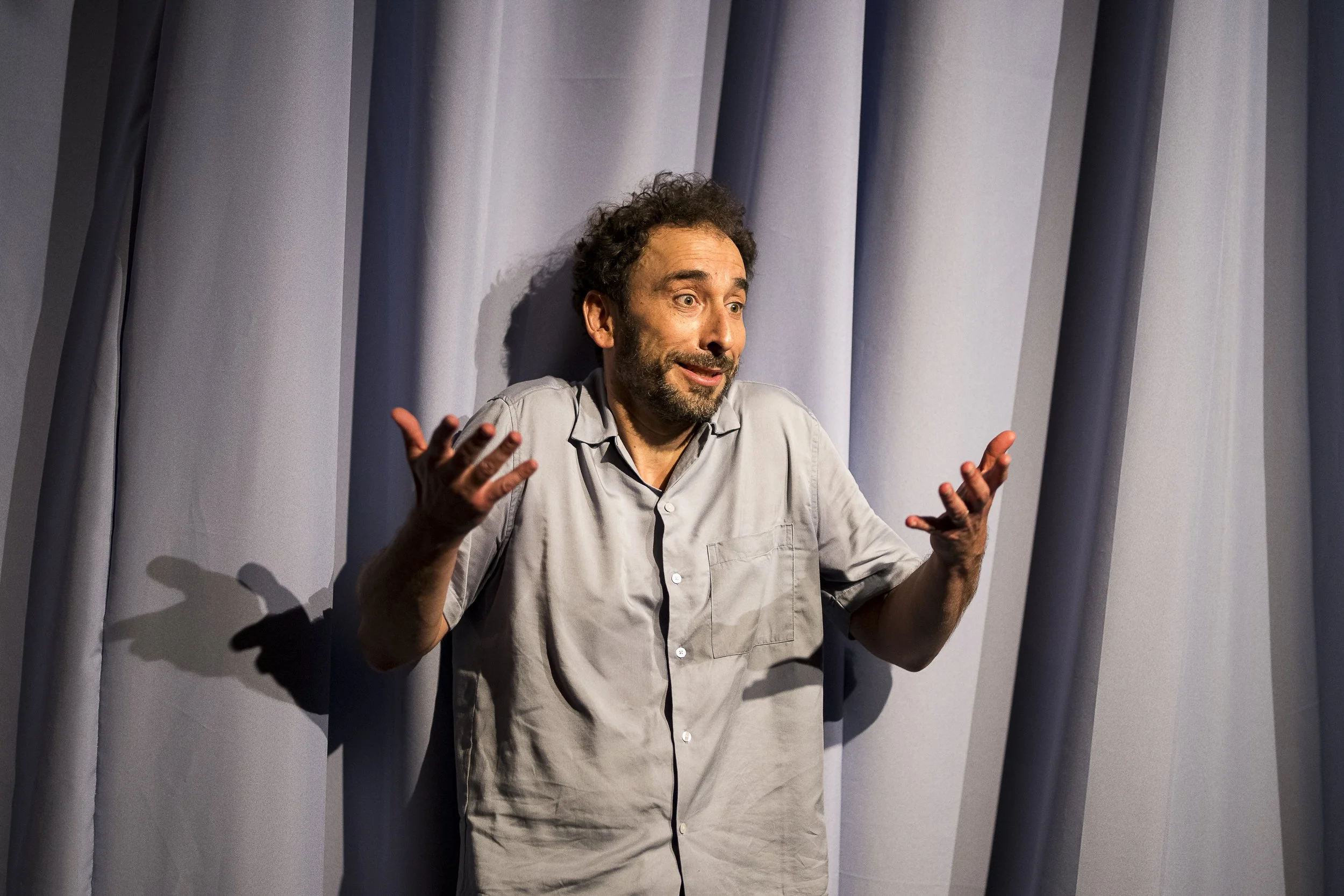 Mann mit verwirrender Gestik vor grauem Vorhang.