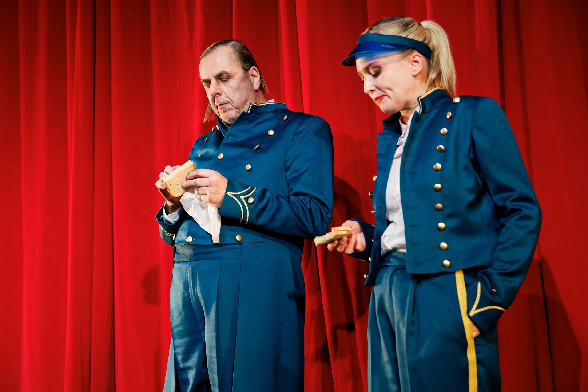 Zwei Schauspieler in blauen Militäruniformen stehen vor roten Bühnenvorhängen und schauen nach unten, während sie Toastbrot essen.