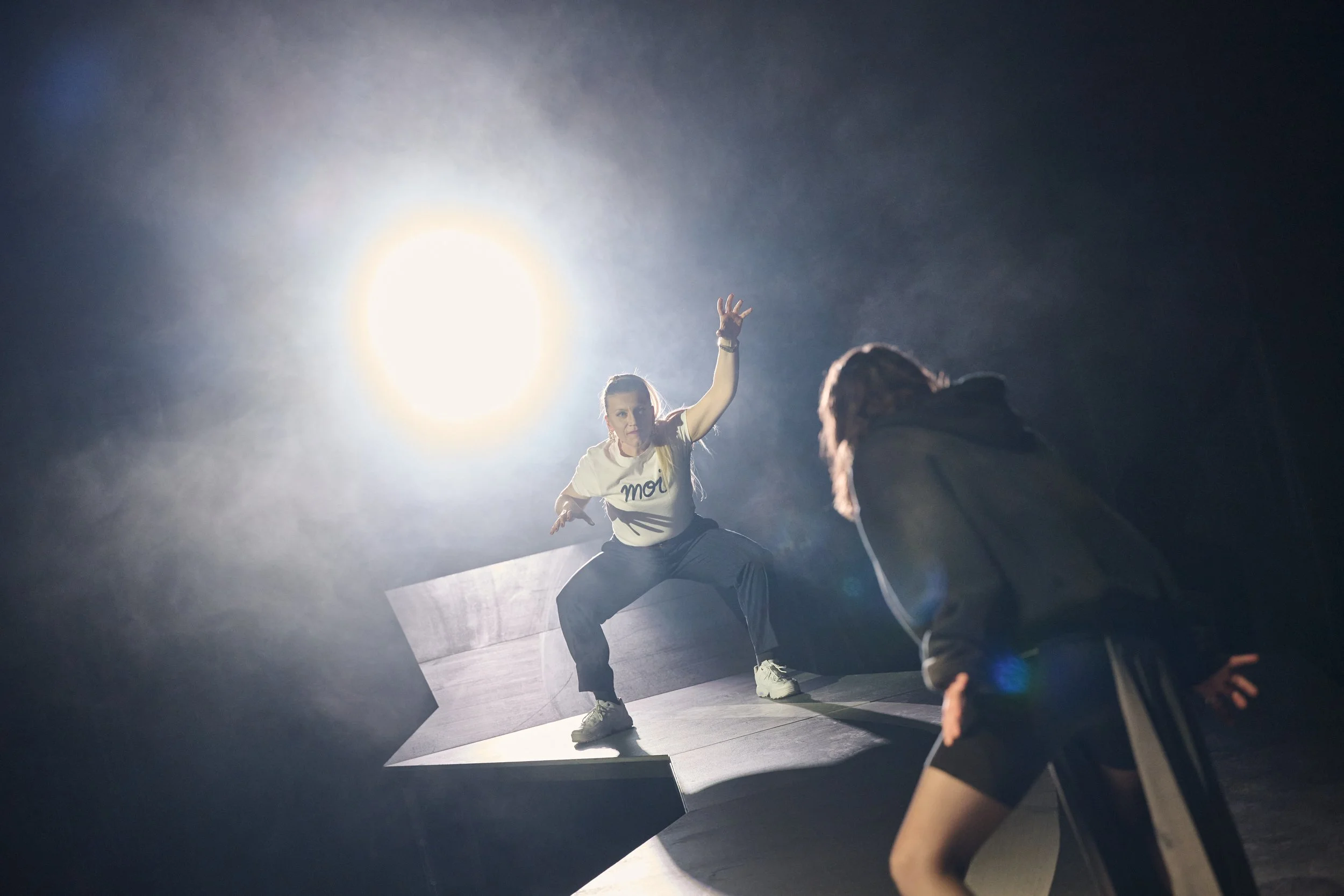 Zwei Frauen bei einer Performance auf einer Bühne, eine davon steht in einer dynamischen Pose, während die andere ihnen gegenübersteht, bei starker Beleuchtung im Hintergrund.