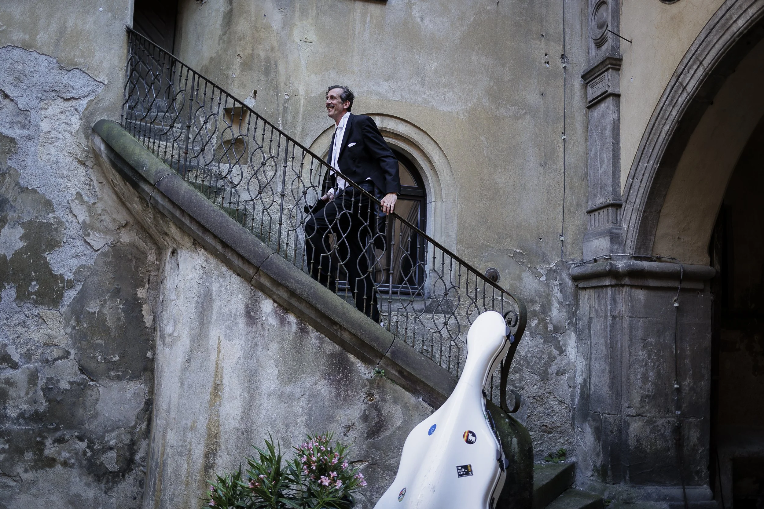 Ein Mann in Anzug, der die Treppe hinauf geht, neben einer weißen Gitarre oder einer ähnlichen tragbaren Lautsprechereinheit, im alten Gebäude mit Steinwänden und einem Fenster.