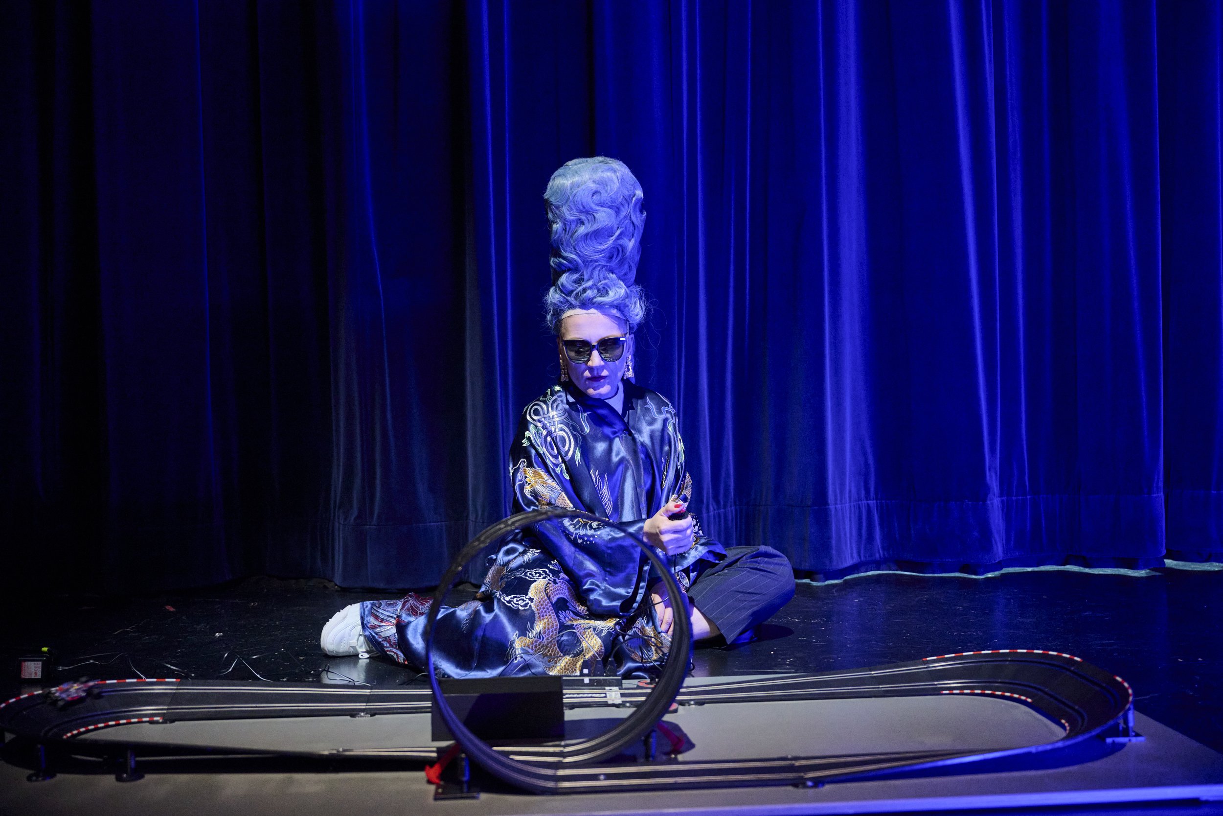 Künstlerin in auffälligem Look mit großer gewellter blauer Frisur und dunkler Sonnenbrille sitzt auf der Bühne vor blauer Vorhang, umgeben von Modellbahn. Stilvolle Kleidung mit goldenen Mustern, große Ohrringe, dunkle Lippen, kontrolliert gestylte H